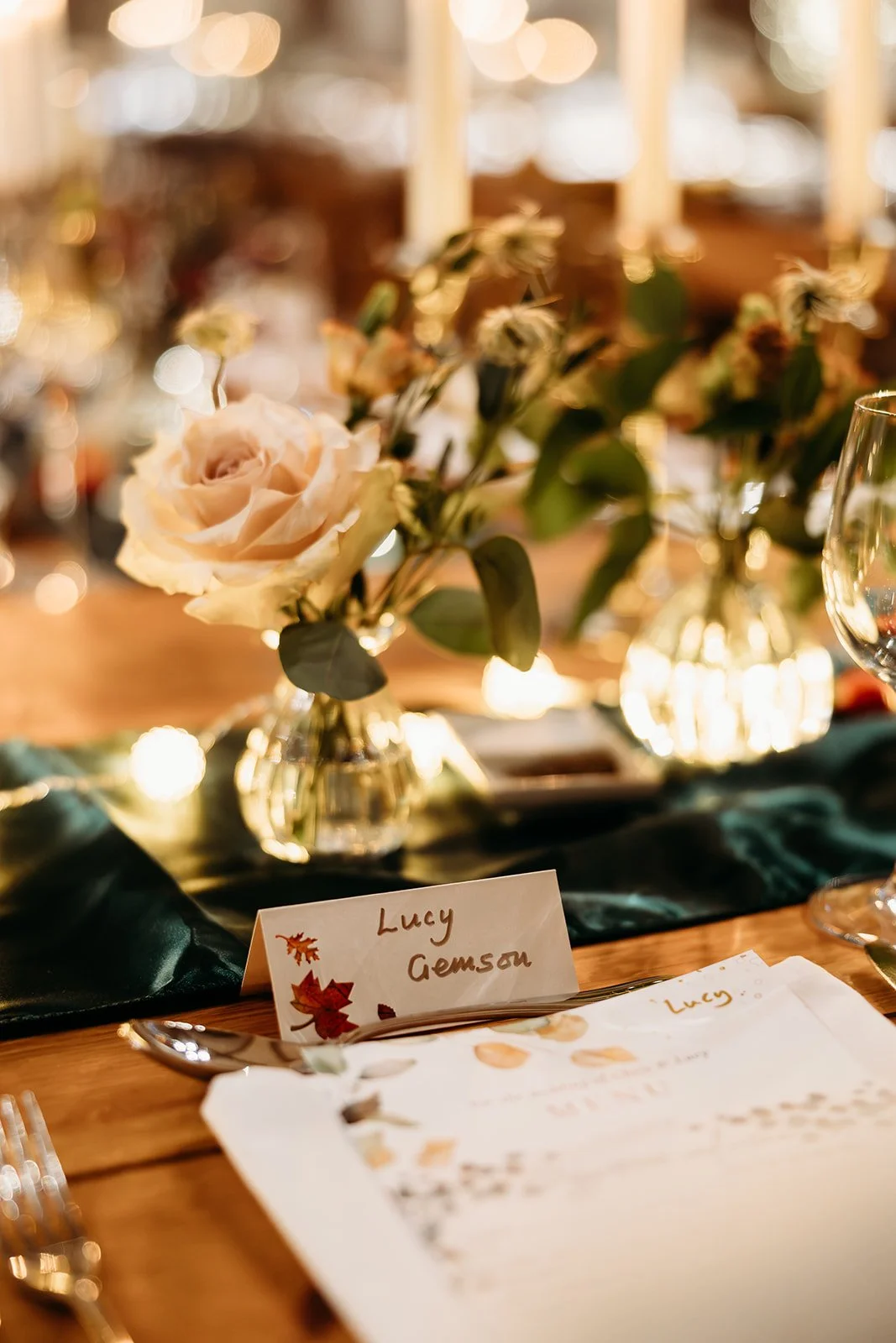 A close-up of a table centerpiece with a beige rose, green leaves, and white flowers in a small glass vase, with a place card reading "Lucy Gemson" beside a menu on a dark table runner, and blurred warm lighting in the background.