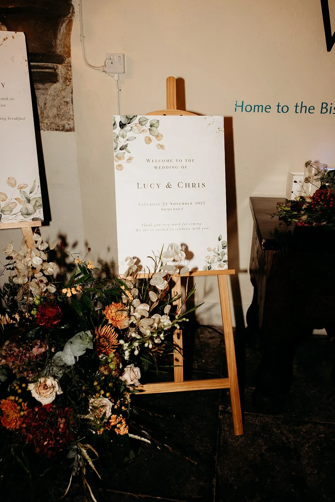 Wedding welcome sign with floral decorations, displayed on a wooden easel, for Lucy and Chris's wedding on November 22, 2025, in Somerset.
