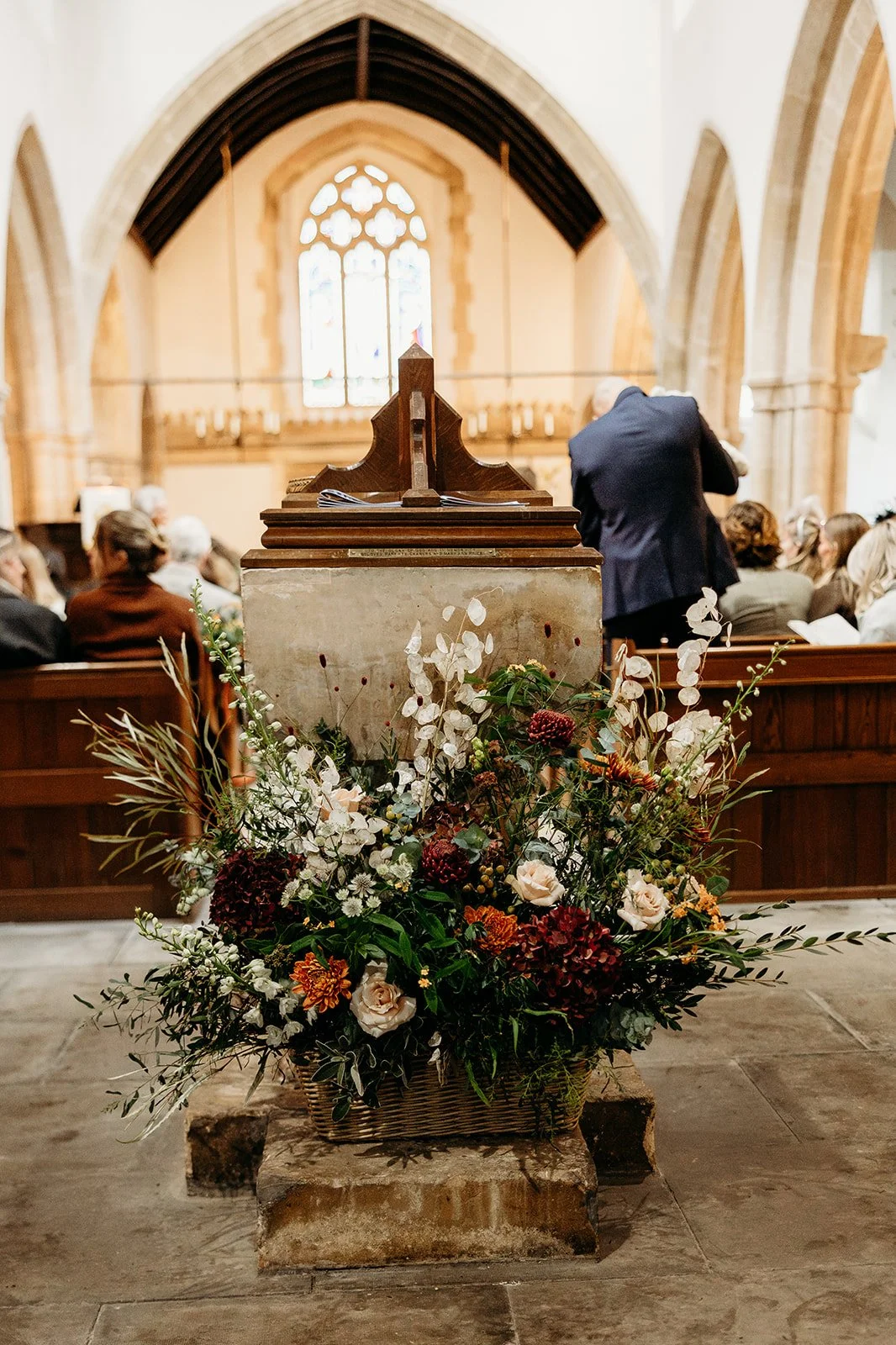 Interior of a church with a large floral arrangement in the foreground, and people seated and standing near the altar in the background, with stained glass windows and Gothic architecture.