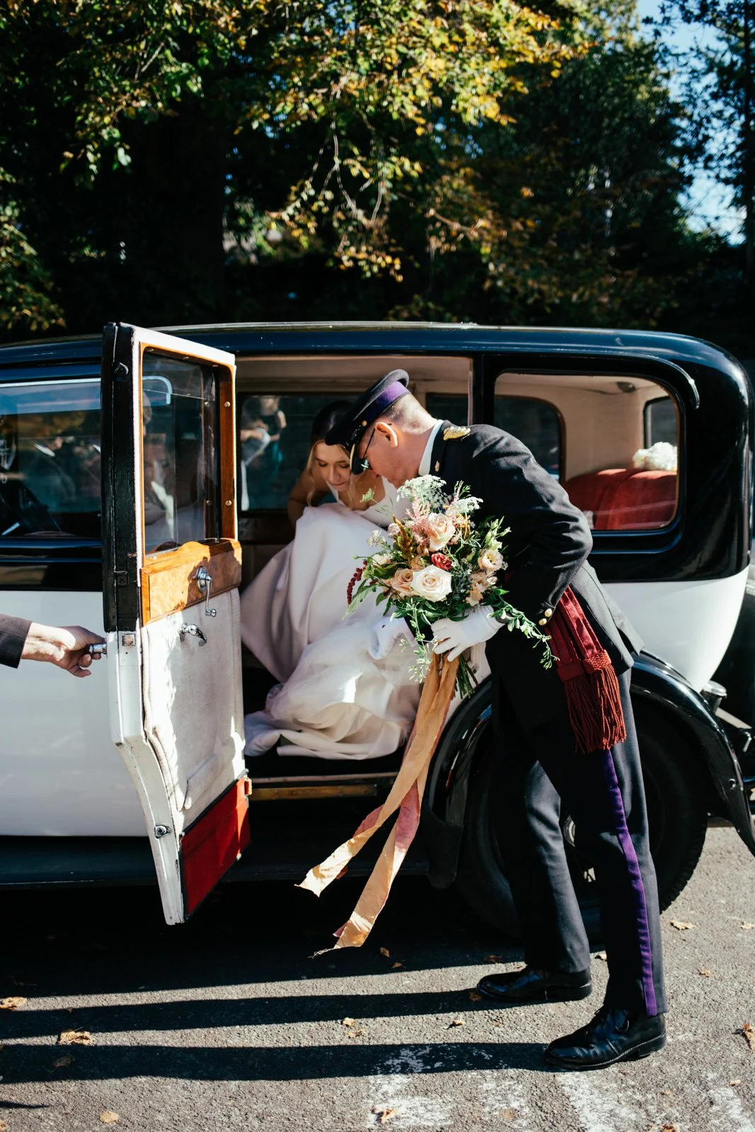 A groom helps a bride out of a vintage black and white car, holding a bouquet of flowers, on her wedding day outside with trees in the background.