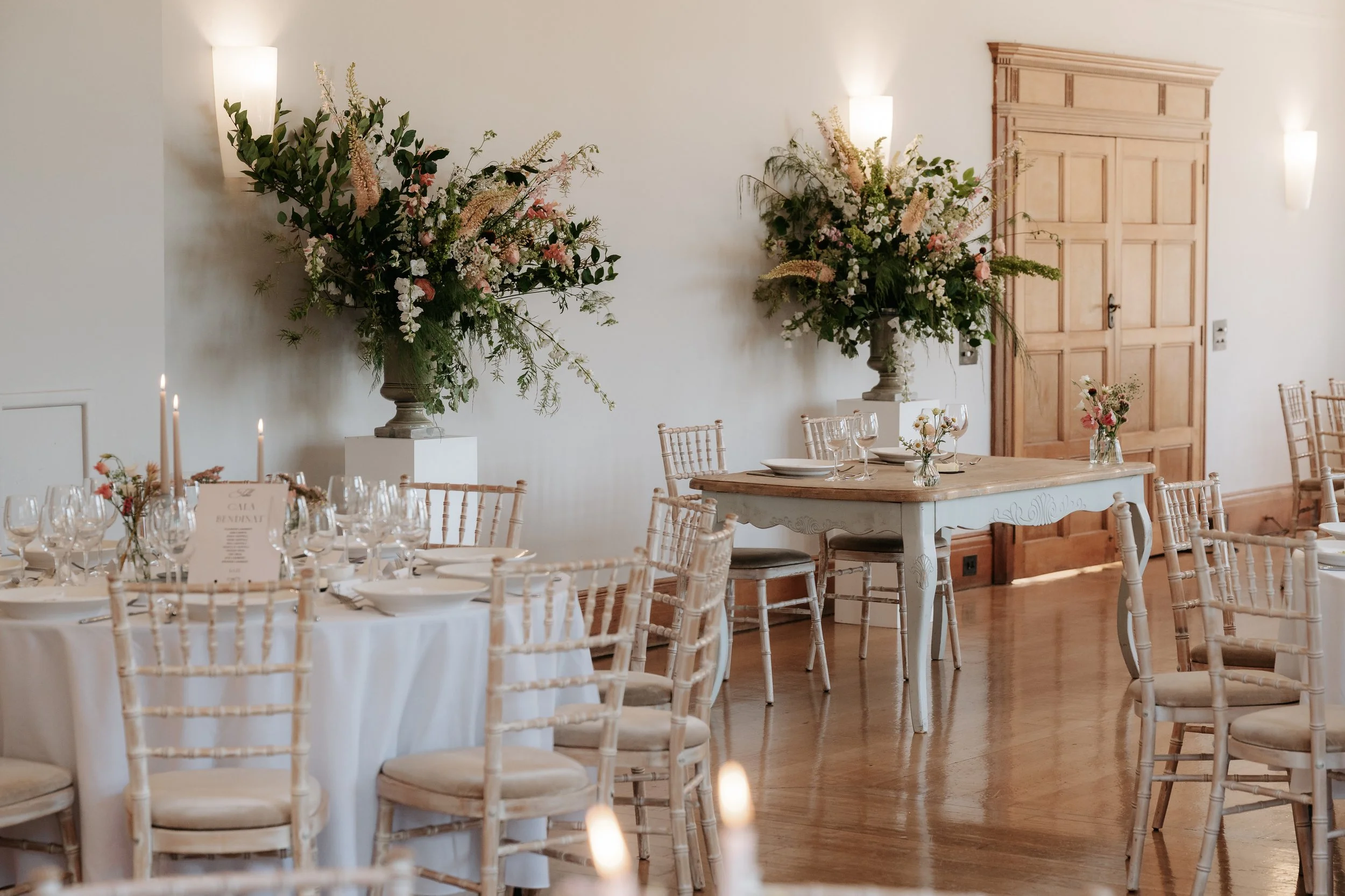 Elegant event space with white round tables, beige chairs, floral centerpieces, and large floral arrangements on tall pedestals, set in a room with white walls, wooden doors, and soft wall-mounted lights.