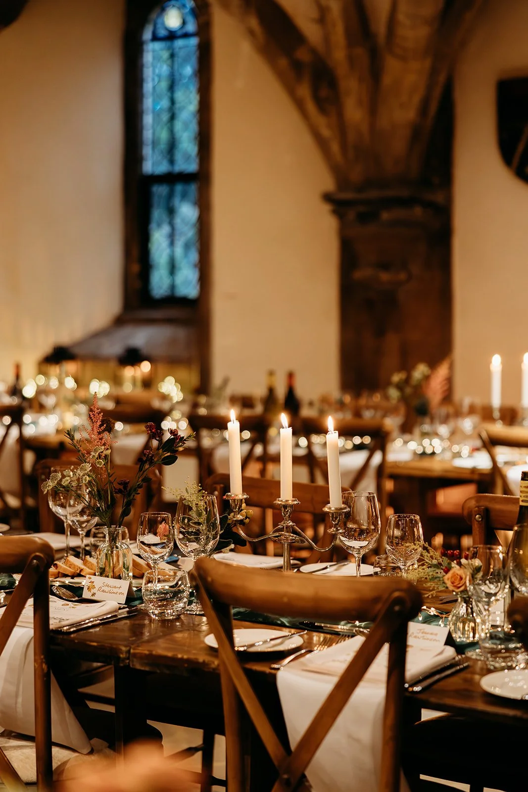Elegant dinner setup with lit candles, glassware, flowers, and place cards in a warmly lit room with wooden beams and stained glass window.