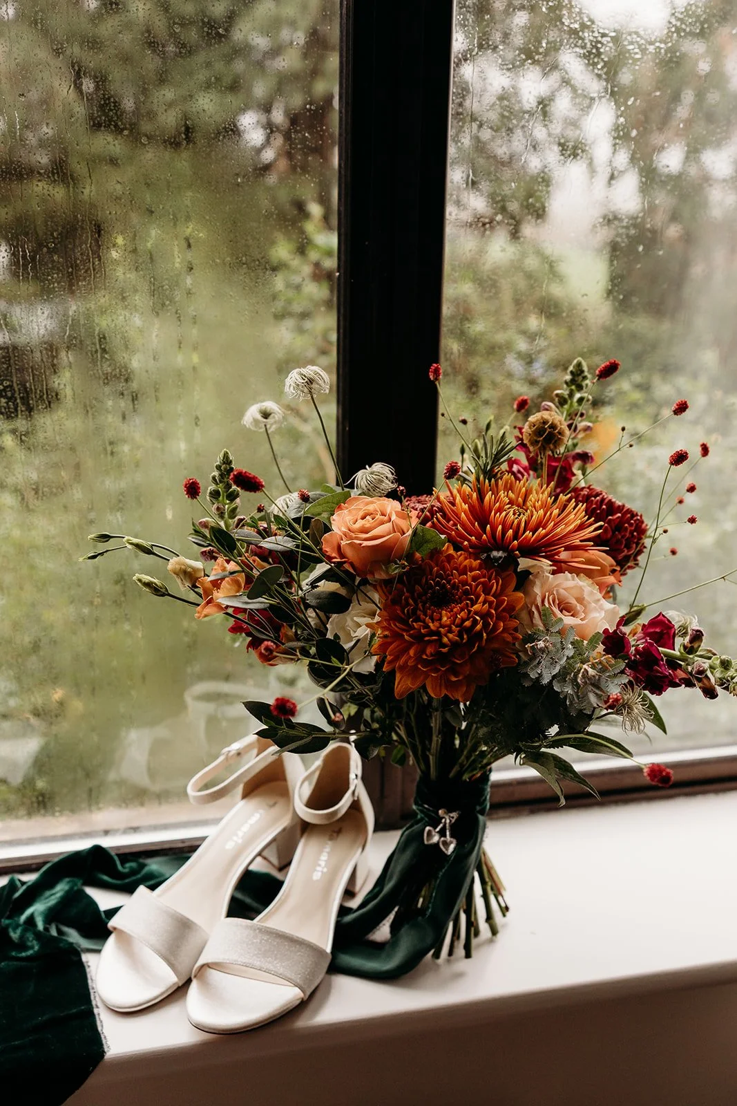 A bouquet of flowers including orange and pink roses, orange chrysanthemums, and other colorful blooms on a windowsill with white high-heeled shoes and a dark green cloth nearby, with rain-streaked window glass in the background.