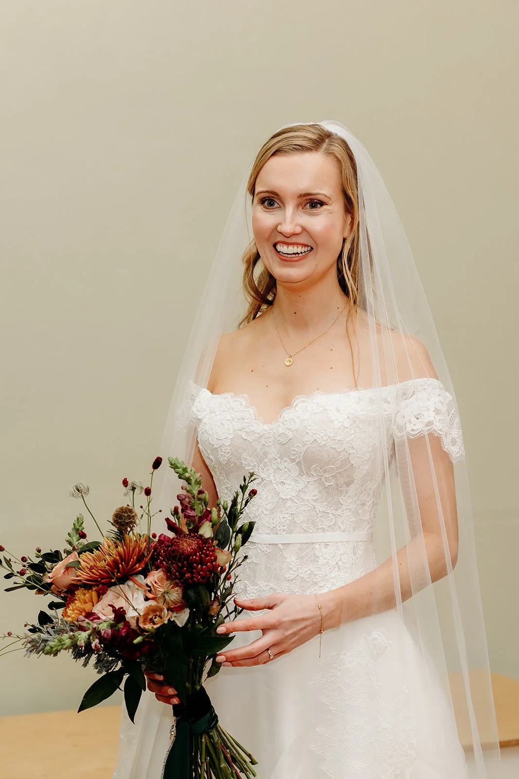 A smiling bride in a white lace wedding dress holding a bouquet of colorful flowers, wearing a veil and a small gold necklace.