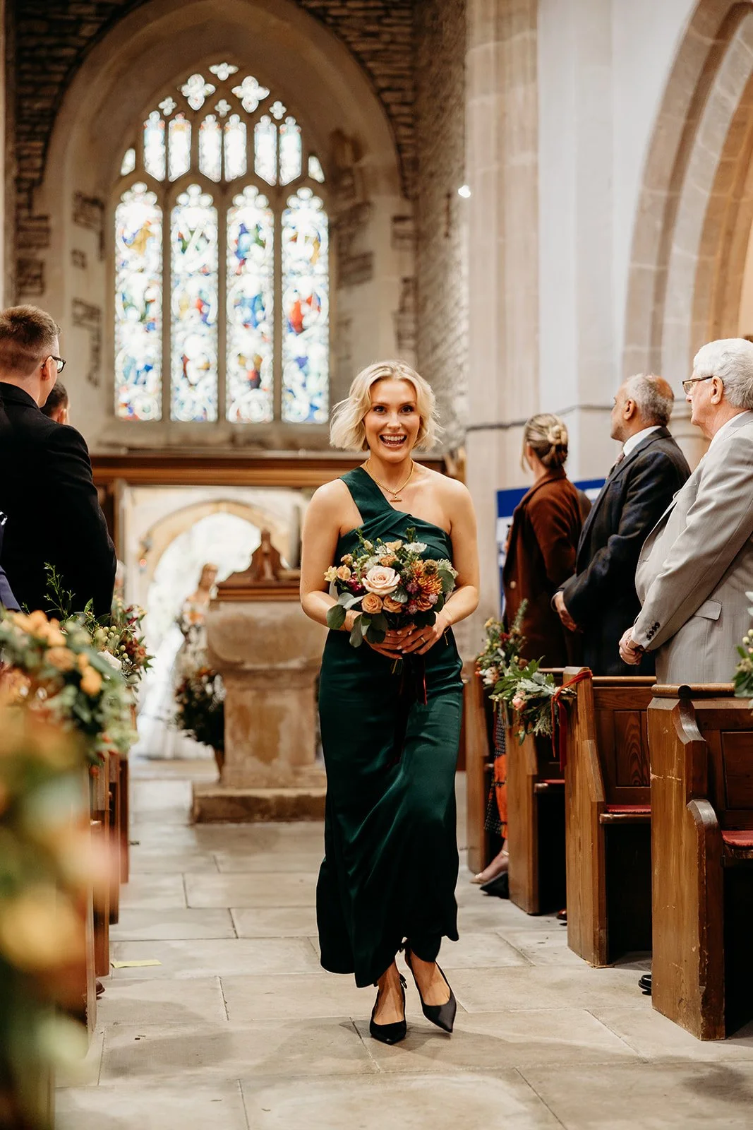 A woman in a dark green dress holding a bouquet walks down the aisle of a church during a wedding ceremony, with people seated on either side and stained glass windows in the background.