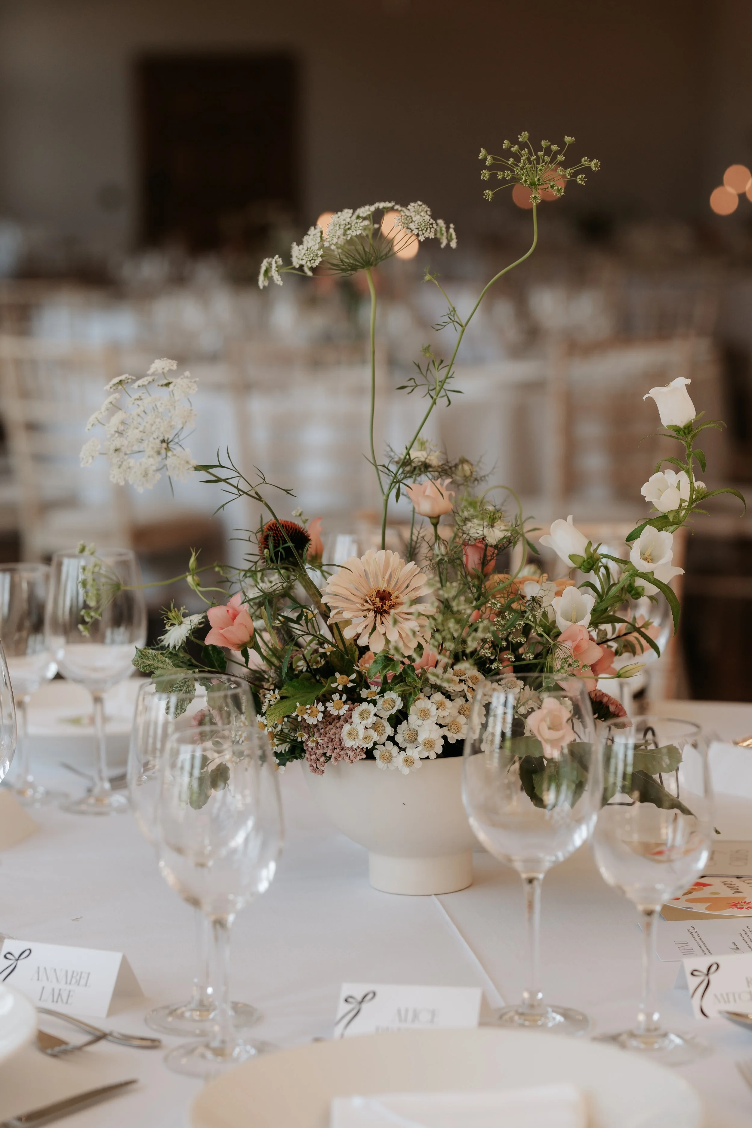 Centerpiece with white, pink, and green flowers on a white tablecloth, surrounded by glasses, cutlery, and place cards at an event.