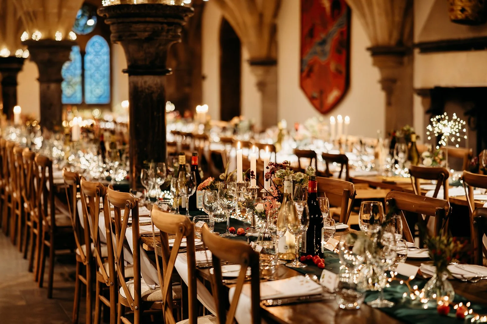 Elegant banquet hall decorated for a formal event or celebration, with long tables set with wine glasses, candles, flowers, and bottles, under warm lighting and gothic architectural features in the background.