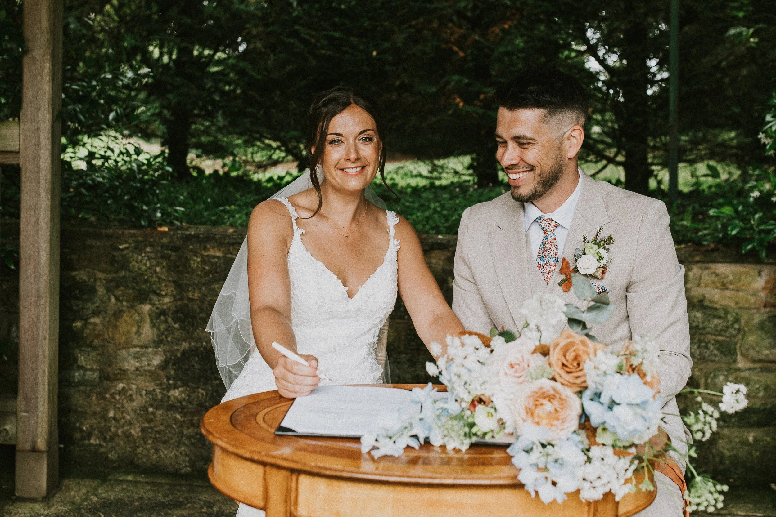 A bride and groom sitting at a table outdoors, signing a document during their wedding ceremony, with a floral arrangement on the table and a green, wooded background.