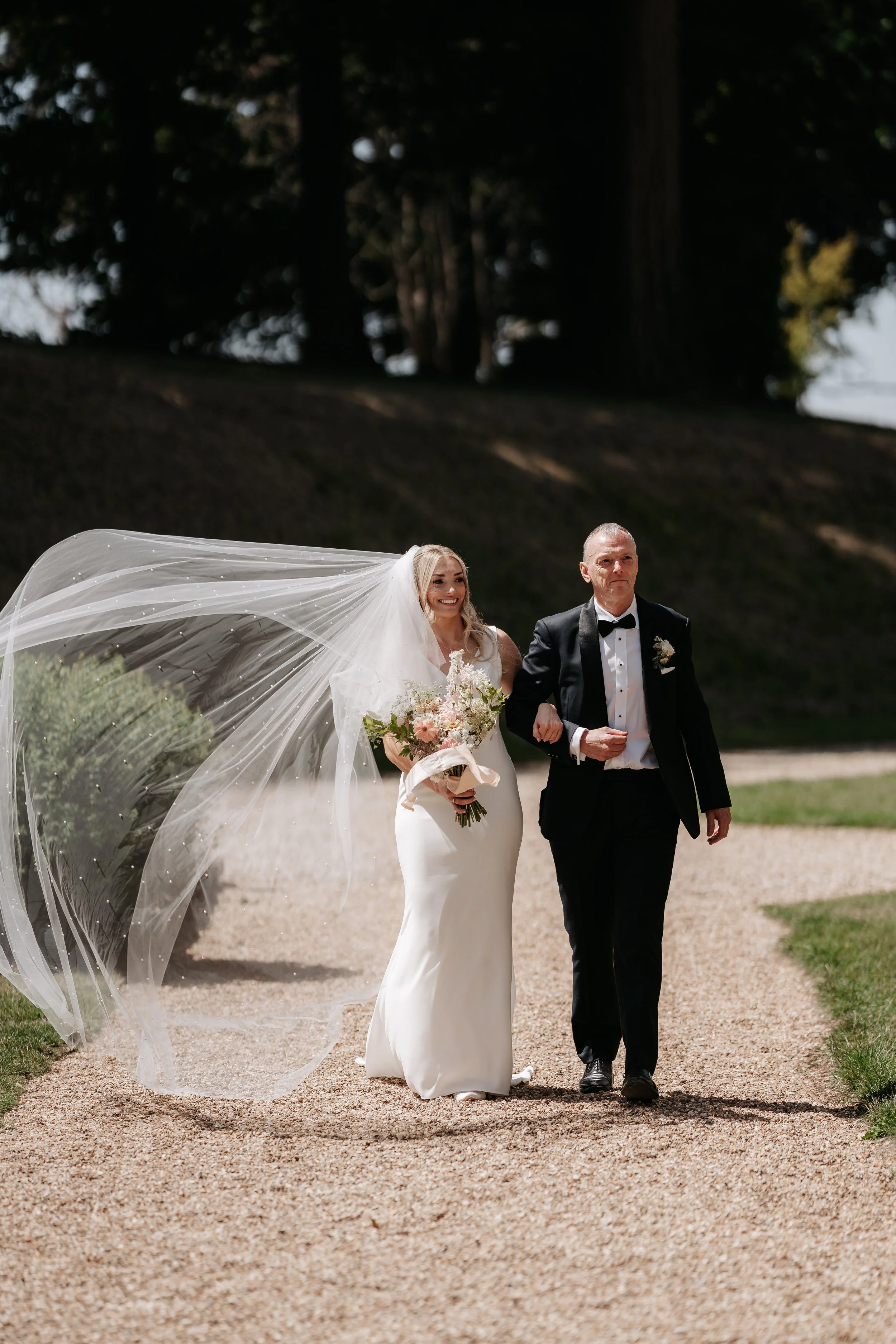 A bride in a white wedding dress and a veil walking beside an older man in a tuxedo, outdoors on a gravel path with trees in the background.