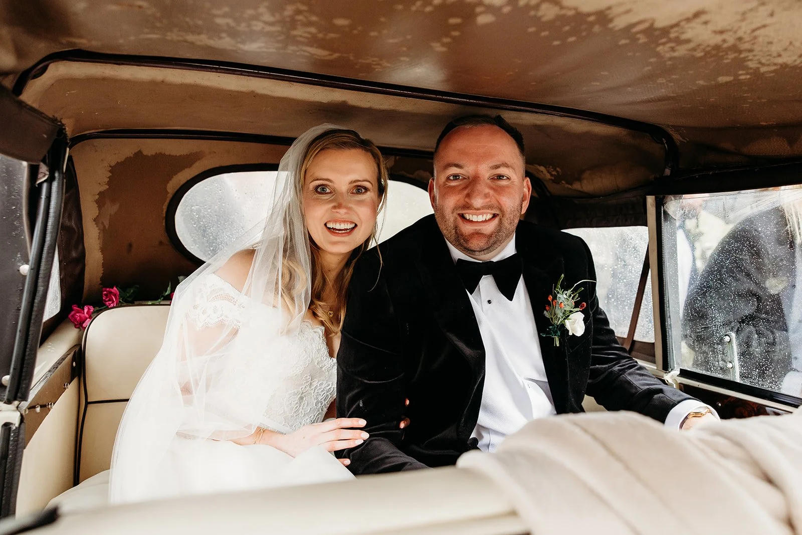 A bride and groom sitting inside a vintage car, smiling at the camera on their wedding day. The bride is wearing a white wedding dress and veil, and the groom is dressed in a black tuxedo with a boutonniere.