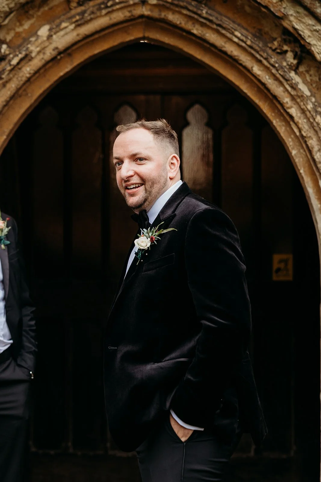 A man in a black tuxedo with a boutonniere, standing in front of a wooden archway with gothic arches, smiling.