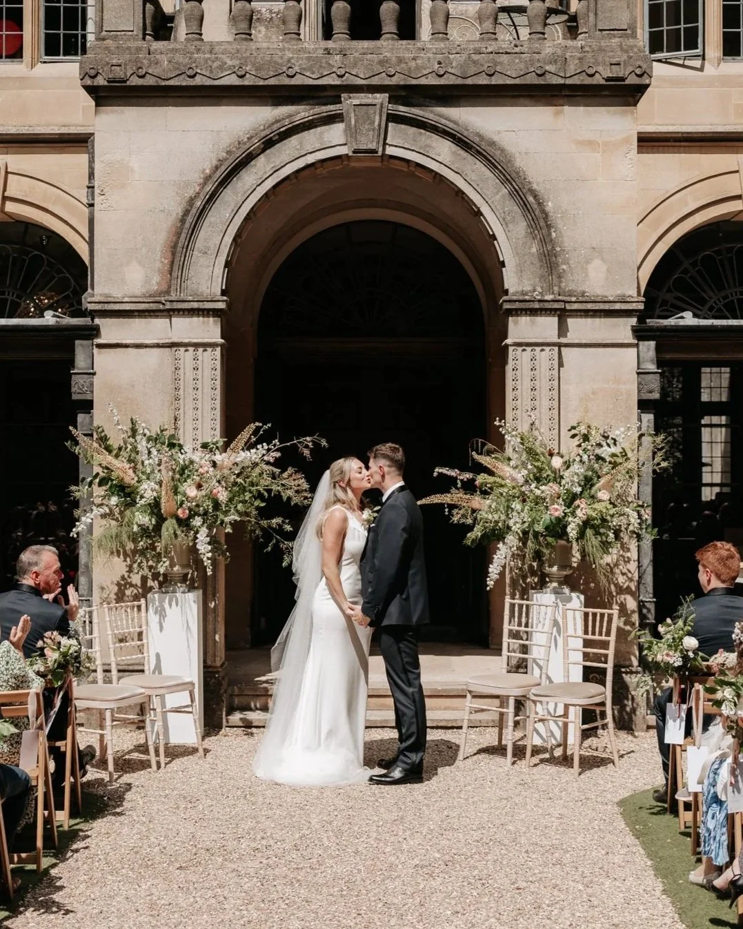 A bride and groom sharing a kiss during their wedding ceremony outdoors, with guests seated around them and floral arrangements on either side.