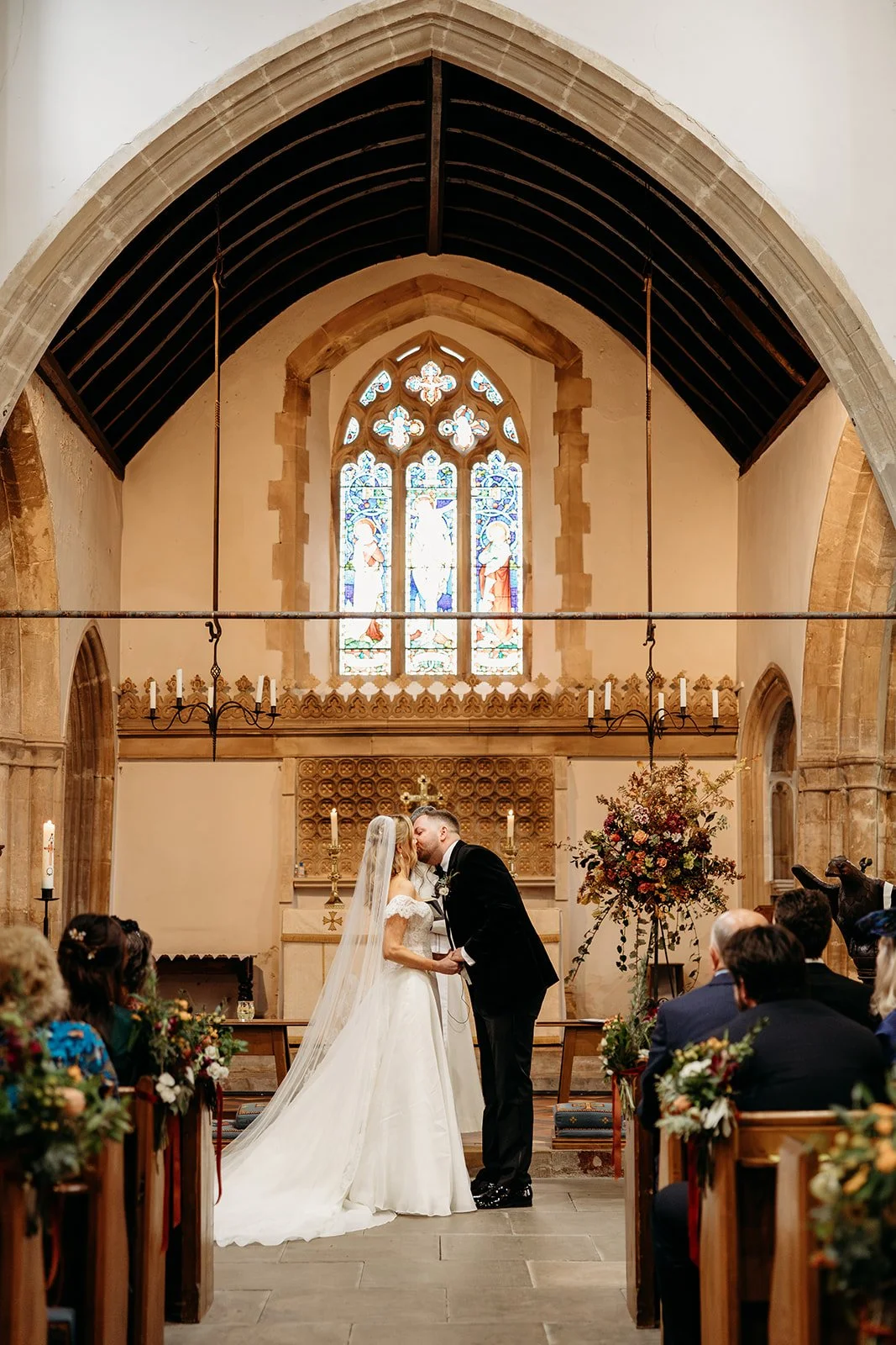 A bride and groom share a kiss during their wedding ceremony inside a church with stained glass windows, floral arrangements, and candles.