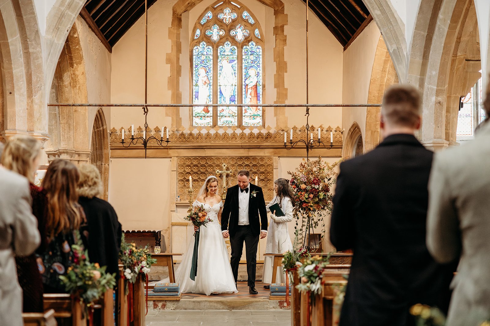 A wedding ceremony taking place inside a church with a bride and groom holding hands at the altar, surrounded by guests. The church has high arched ceilings, stained glass windows, and floral decorations.