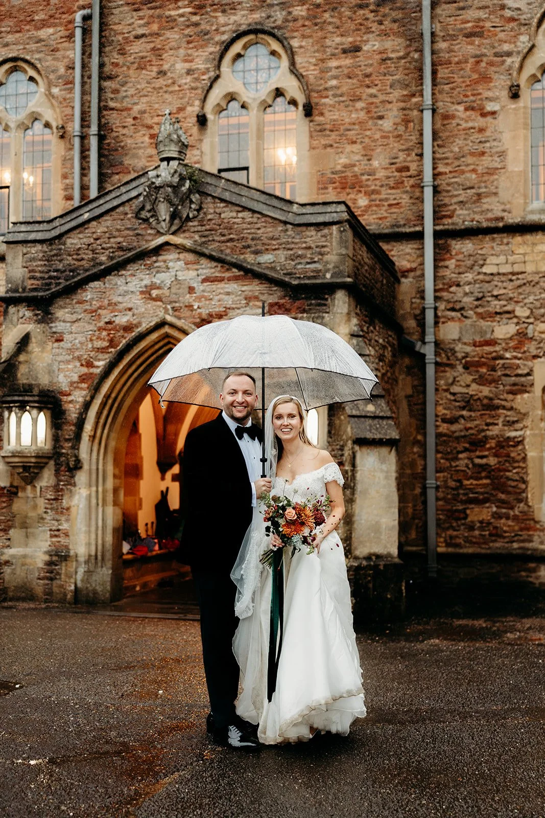 A newlywed couple stands smiling under a transparent umbrella in front of a historic brick church during rainy weather, with the bride holding a bouquet of flowers.