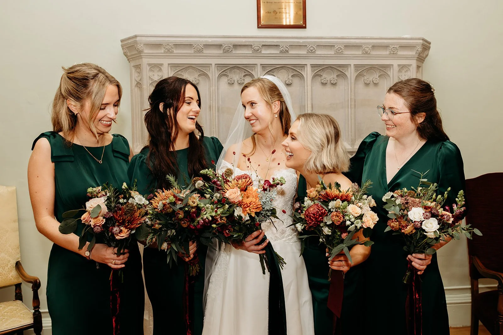 A bride with four bridesmaids, all holding bouquets of flowers, smiling and looking at each other in an indoor setting with a decorative fireplace mantel in the background.