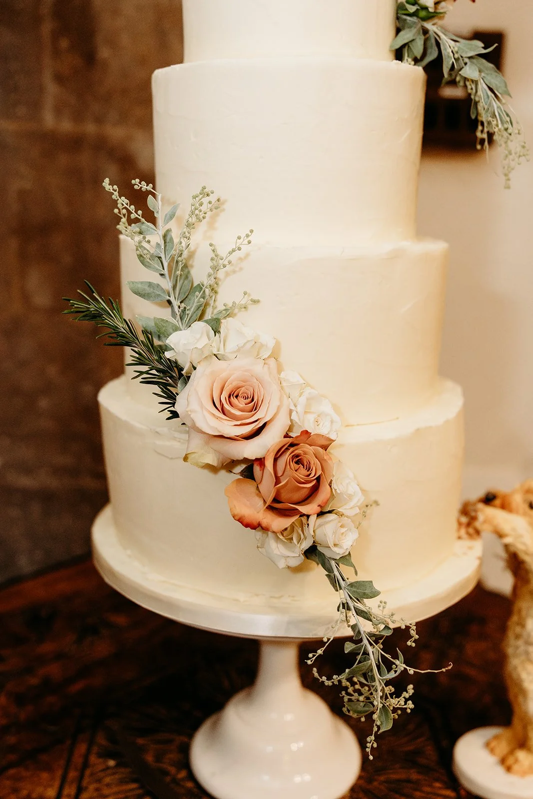 Three-tiered white wedding cake decorated with peach and white roses, green eucalyptus, and other greenery, on a white cake stand.