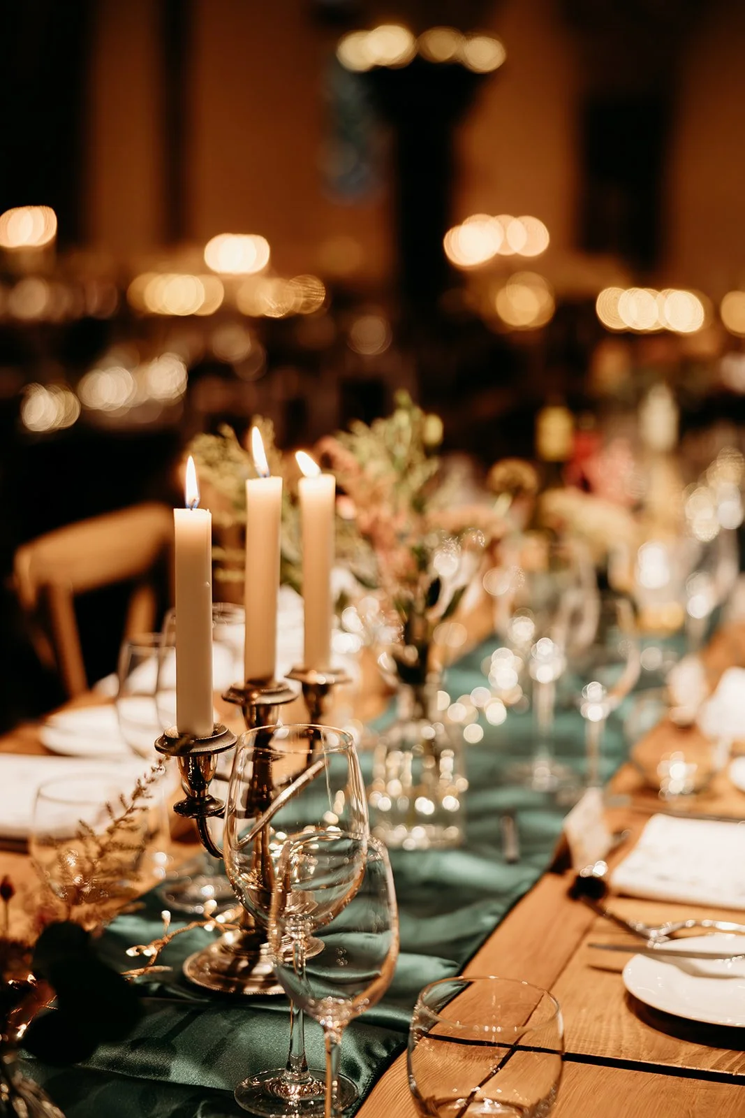A close-up of a candlelit dinner table with three tall white candles in silver candlesticks as the centerpiece, surrounded by wine glasses, plates, and floral arrangements, set in a warmly lit indoor space.