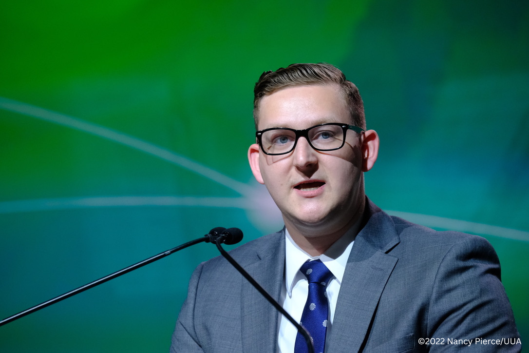 John in a white shirt, blue tie, and gray jacket speaking at a podium. Photo credit watermark reads "Copyright 2022 Nancy Pierce UUA."