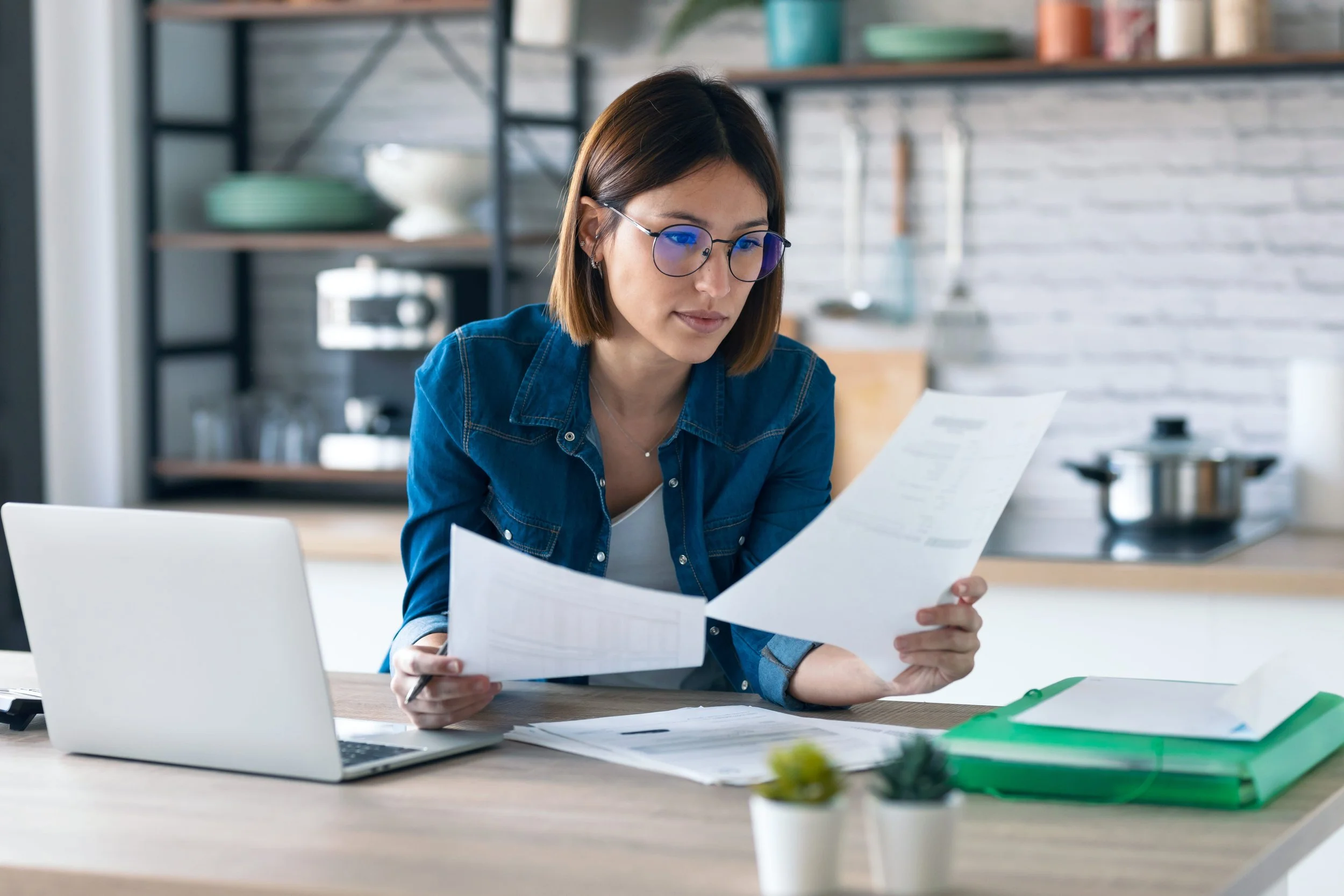 A female accountant reviews financial documents at her desk with a laptop open beside her.