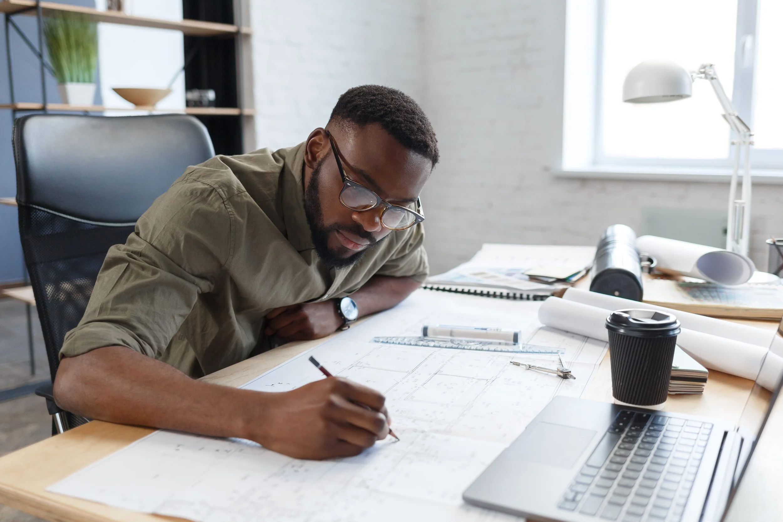 A male architect in glasses works carefully on building plans at his desk with drafting tools and a laptop nearby.