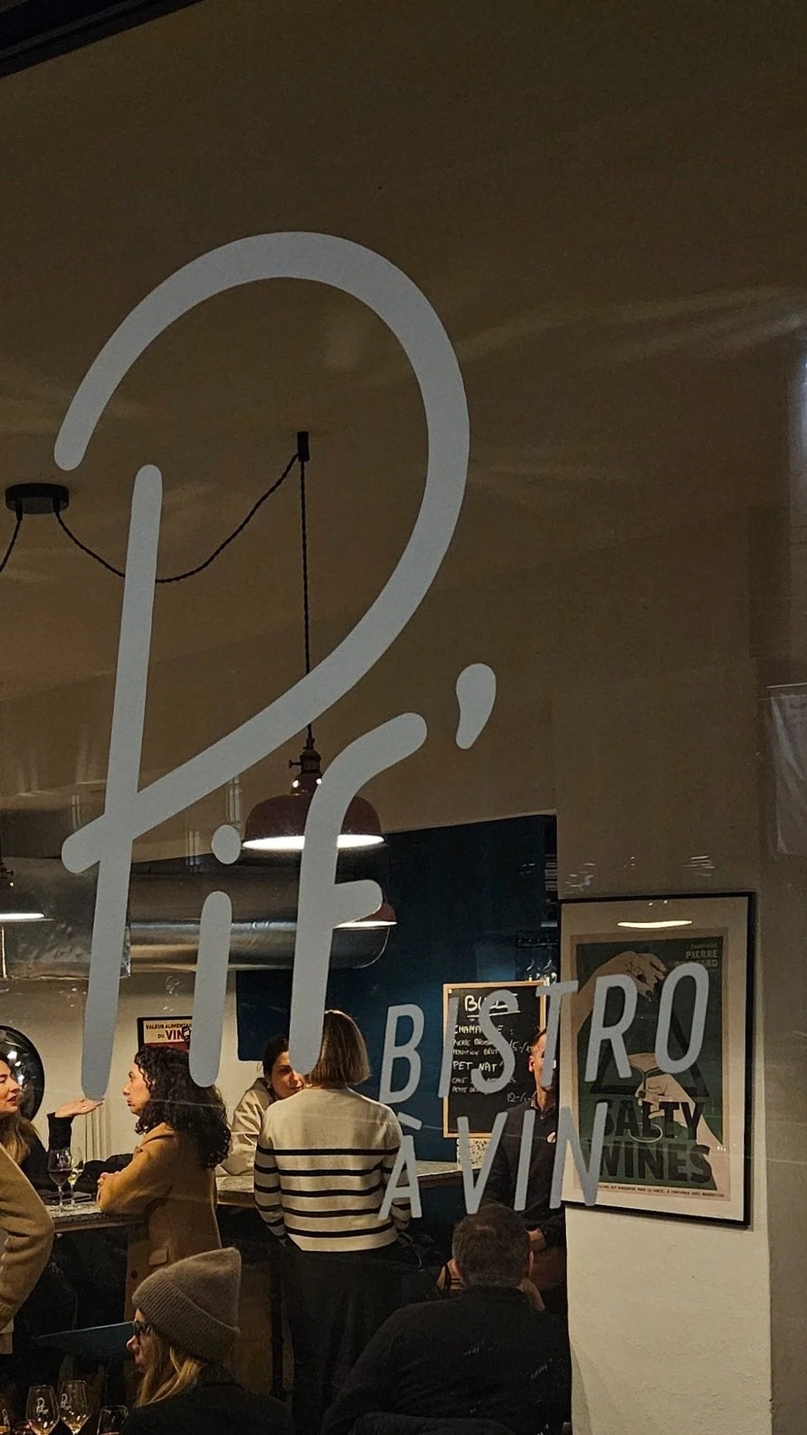 People in a cozy wine bistro with a large logo on the window, engaging in conversation around a table, with wine glasses visible. A poster and menu board are on the wall inside.