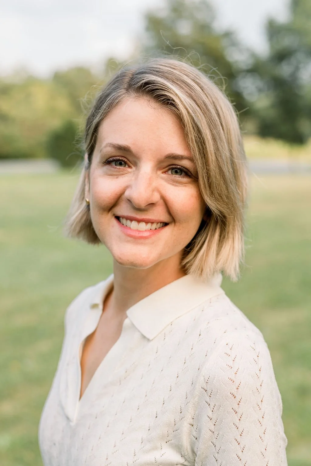 Smiling woman outdoors with short blonde hair and white shirt, standing in a grassy area with blurred trees in the background.