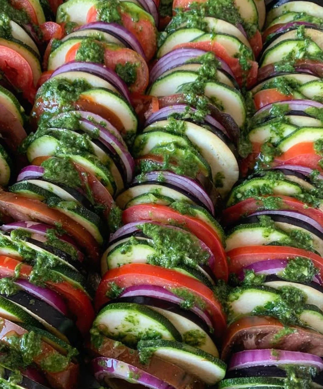 Colorful vegetable ratatouille with slices of zucchini, tomato, onion, and eggplant topped with green pesto sauce.