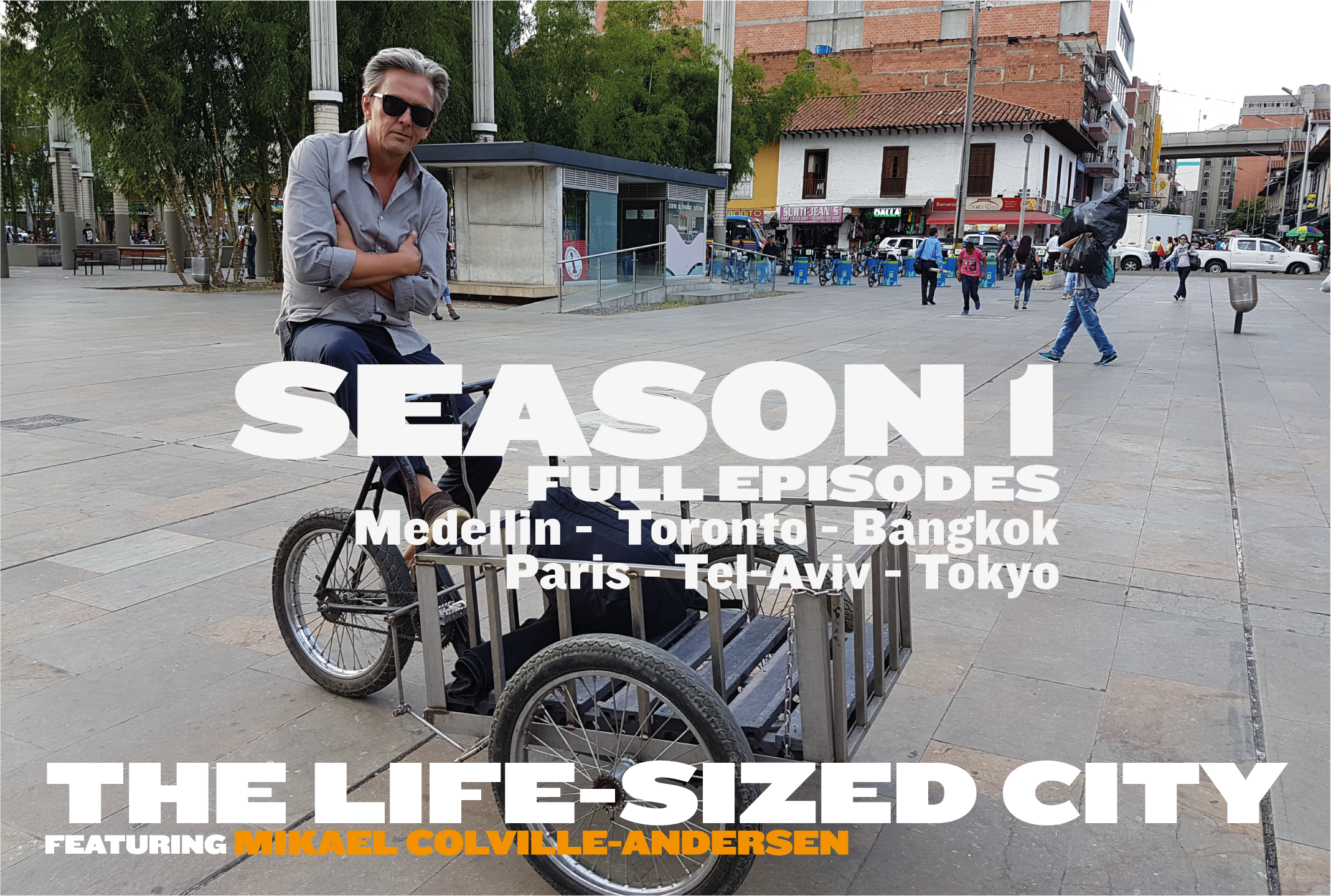 Man in sunglasses sitting on a bicycle with a cart in a city square, with other people walking and buildings in the background.