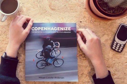 Person holding a book titled "Copenhagenize: The definitive guide to global bicycle urbanism" on a wooden table with coffee, a potted plant, and a cell phone.
