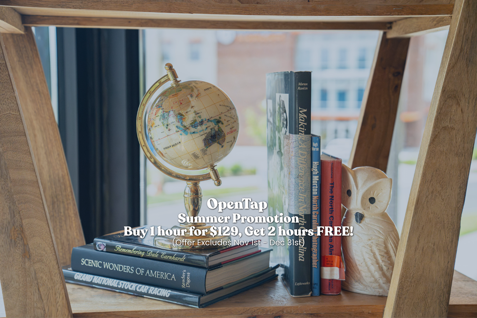 A wooden shelf with a small globe, a stack of books including 'Remembering Dale Earnhardt,' 'Scenic Wonders of America,' and 'Grand National Stock Car Racing,' and a decorative owl figurine in front of a window.
