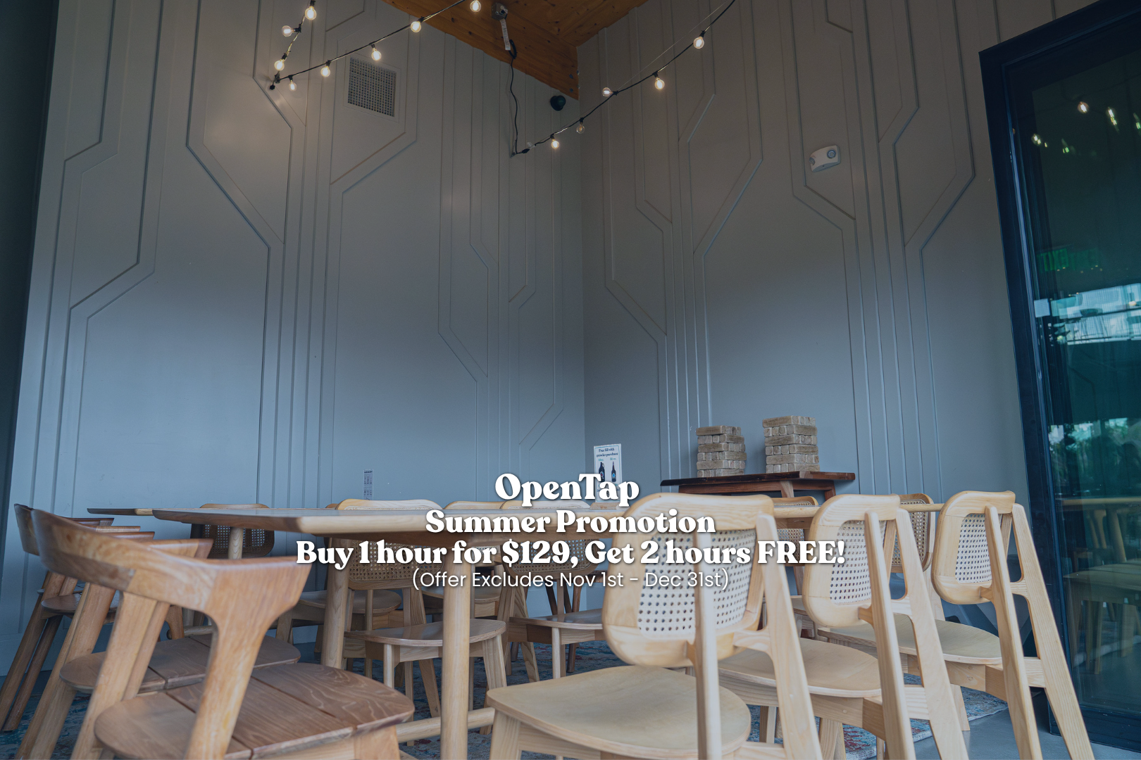 Interior of a dining area with light wooden chairs and a table, a gray wall with geometric patterns, string lights hanging from the wooden ceiling, and a large window.