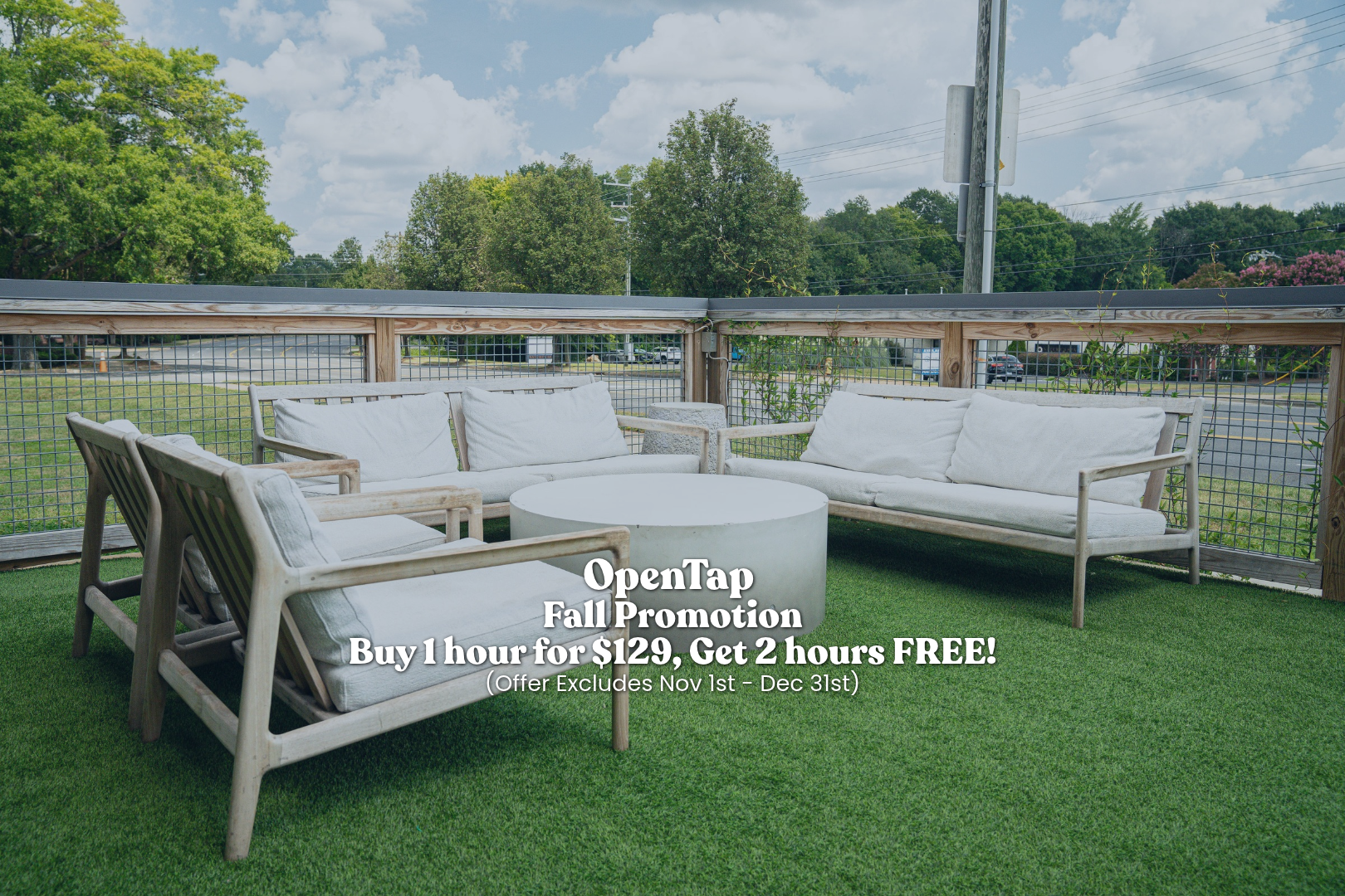 Outdoor patio area with white cushioned seating including a sofa, two armchairs, and a round white table on artificial grass, with trees and a parking lot in the background.