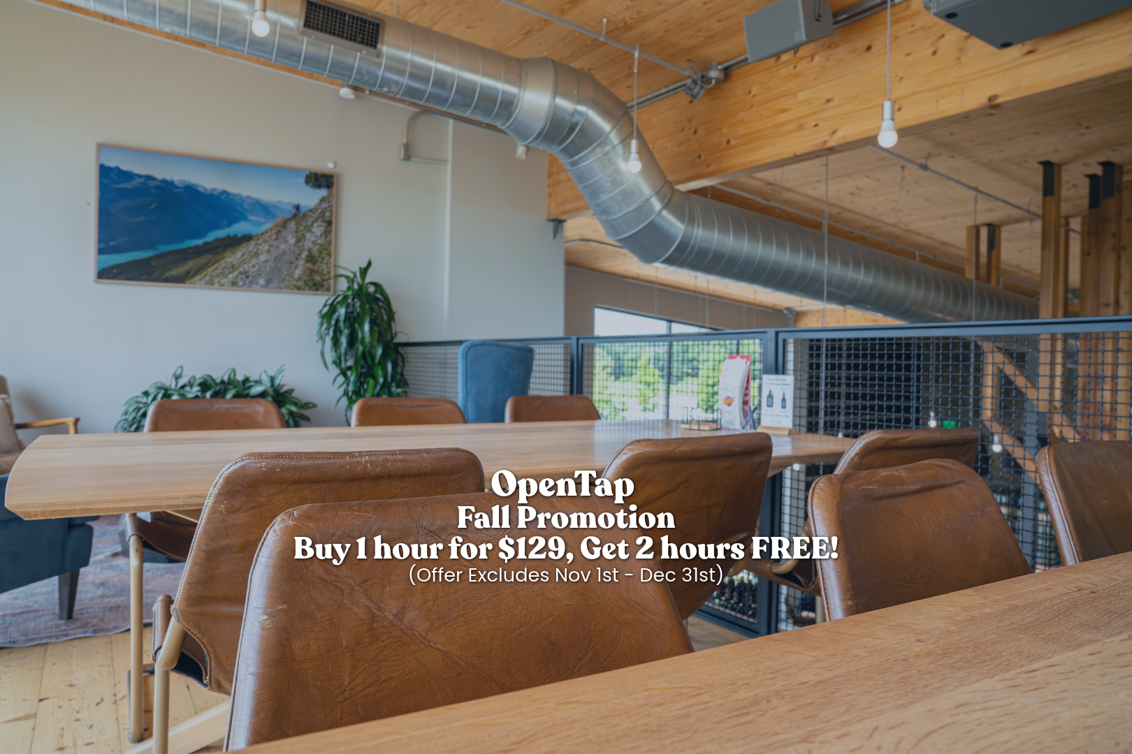 Interior of a modern, airy cafe or meeting space with wooden ceilings, leather chairs around a large wooden table, a large landscape photograph on the wall, and an extensive air duct with pendant lights.