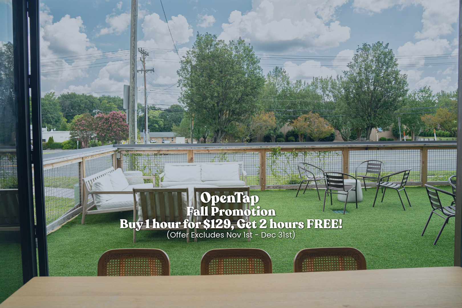 Outdoor patio with artificial grass, seating area with white couches and black metal chairs, view of trees and power lines, partly cloudy sky.
