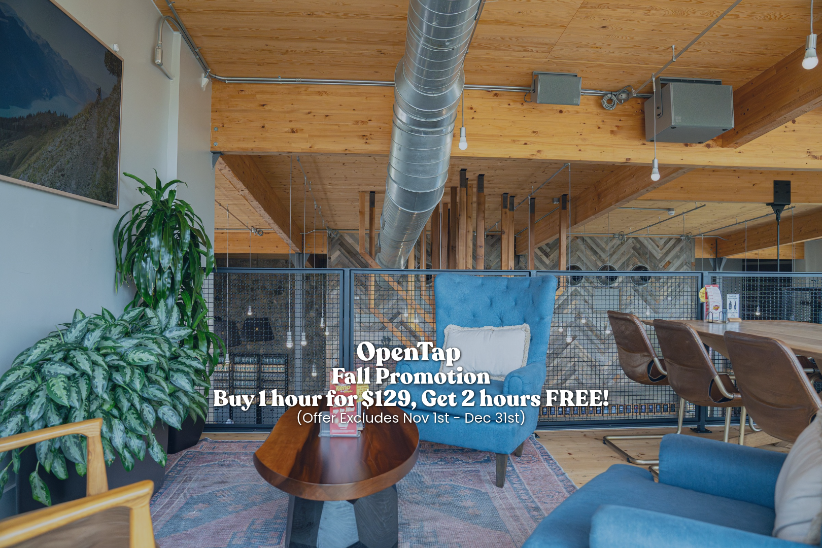 Indoor lounge area with wooden ceiling, green plants, a blue armchair, a wooden coffee table, and a dining table with brown chairs, overlooking a mezzanine with industrial ductwork.