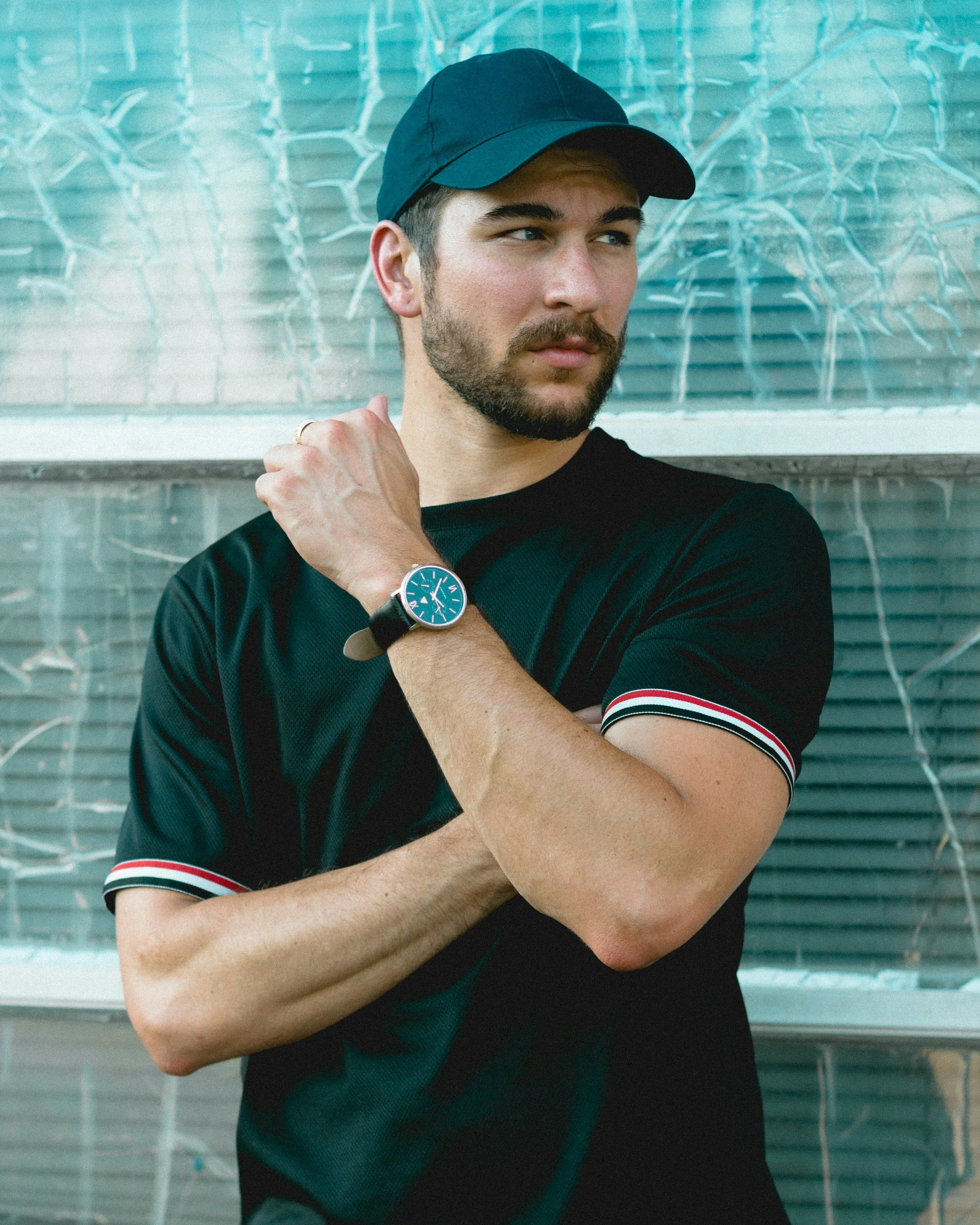 A young man with a beard wearing a blue baseball cap, black t-shirt with red and white stripes on the sleeve, and a wristwatch, standing in front of a textured glass wall.