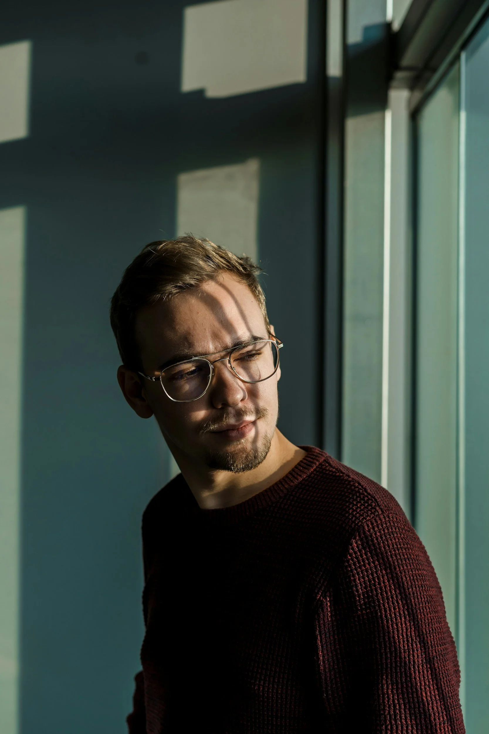 Young man with glasses in a dark red sweater stands near a window, with sunlight casting shadows on the wall.