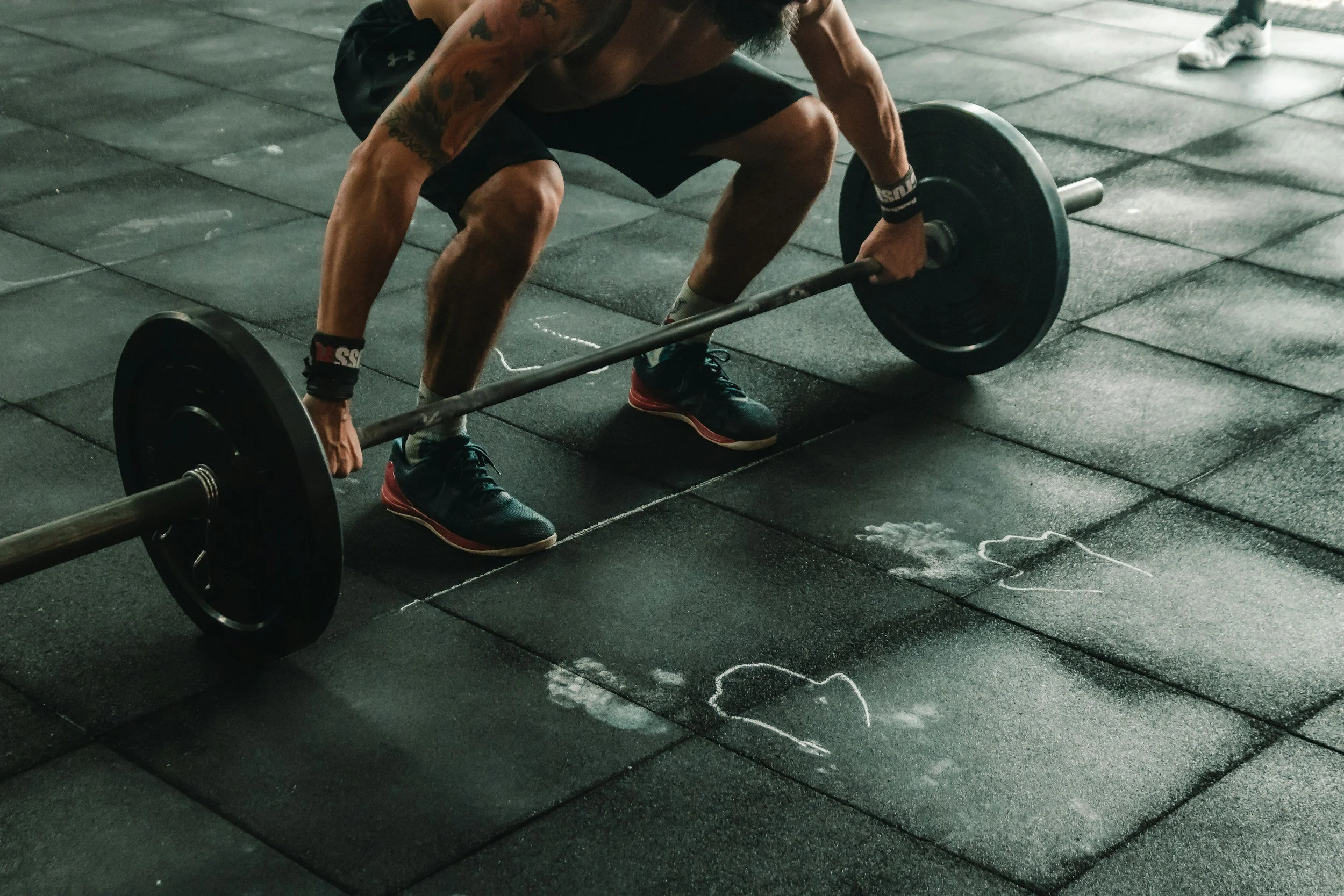 A man with tattoos is lifting a barbell in a gym. He is wearing black shorts, black shoes, and wrist wraps. The gym floor has black rubber tiles with chalk marks on them, and another person's foot is visible in the background.