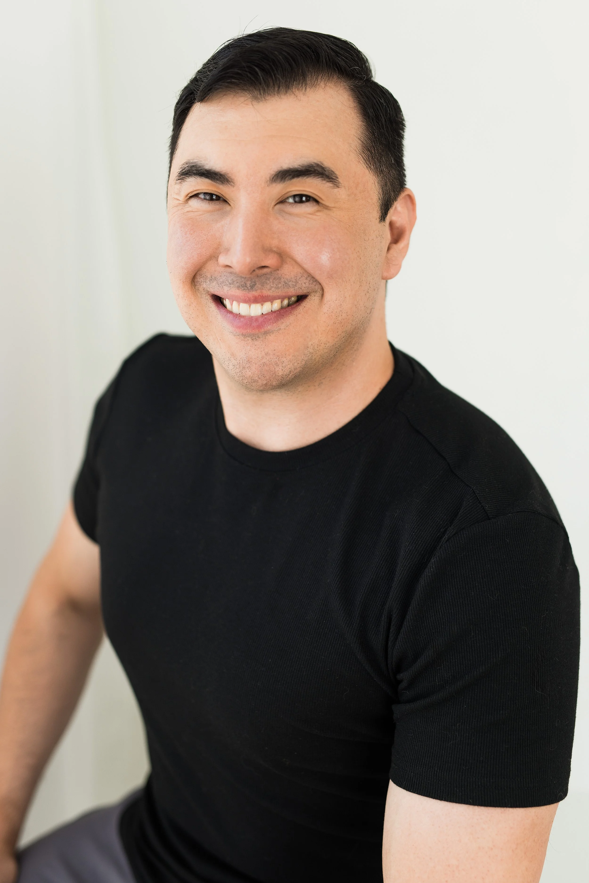 A smiling man with short dark hair wearing a black T-shirt against a plain white wall.