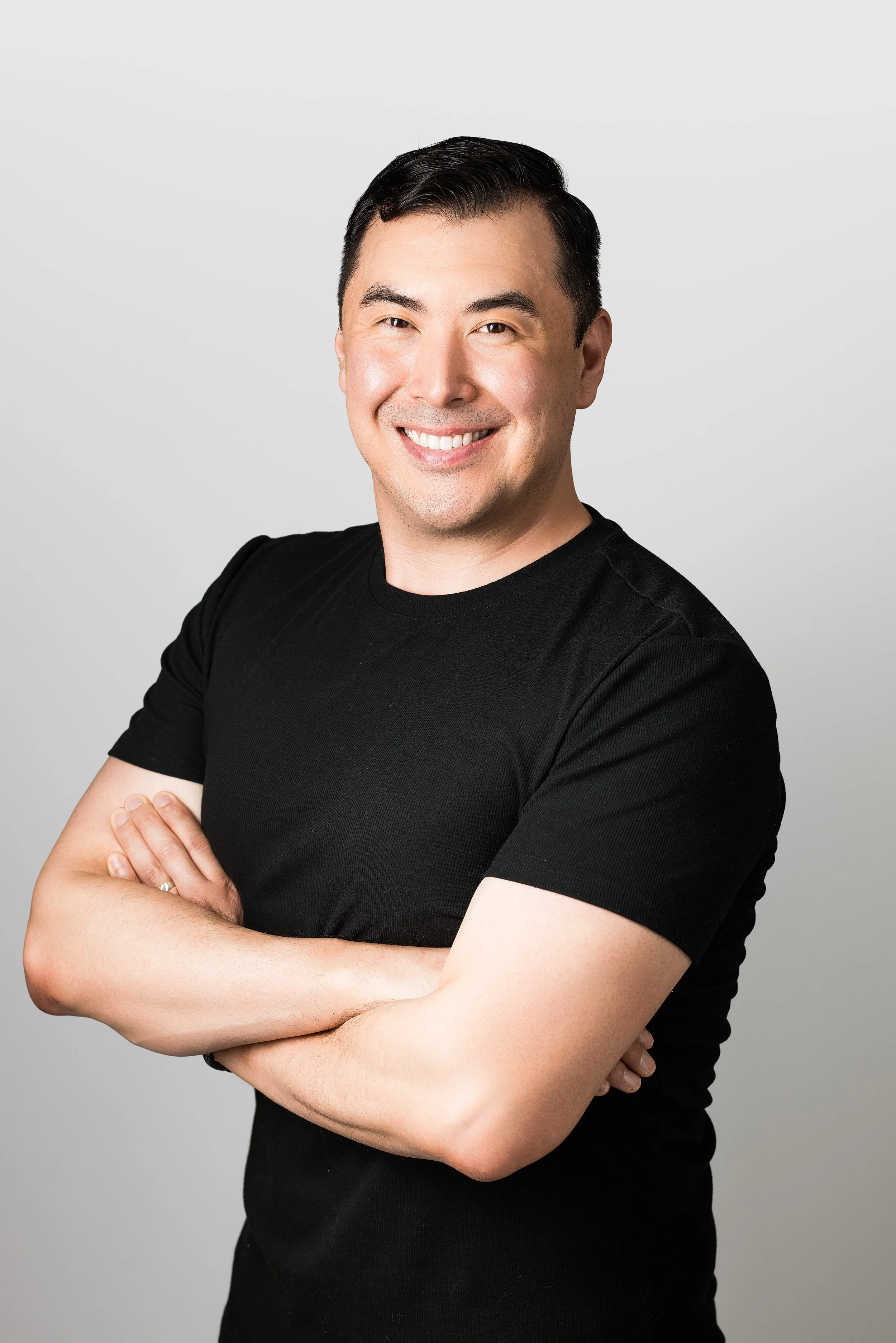 A smiling man with short dark hair, wearing a black t-shirt, standing with arms crossed against a plain light gray background.