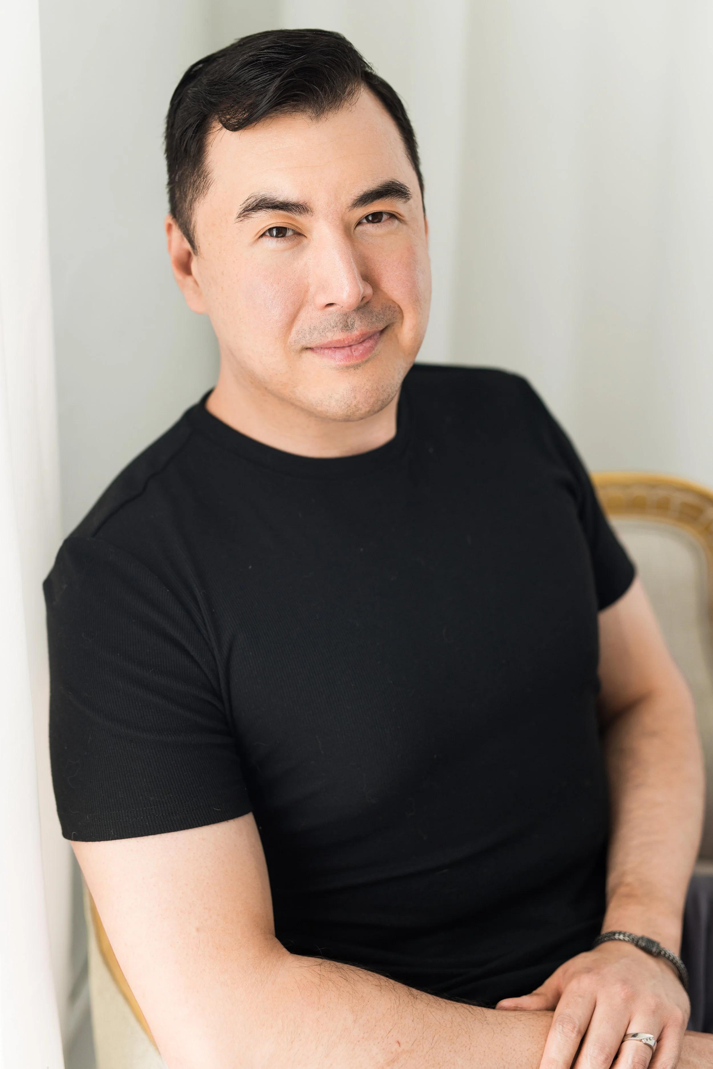A man in a black t-shirt sitting against a white wall, smiling with a neutral background.