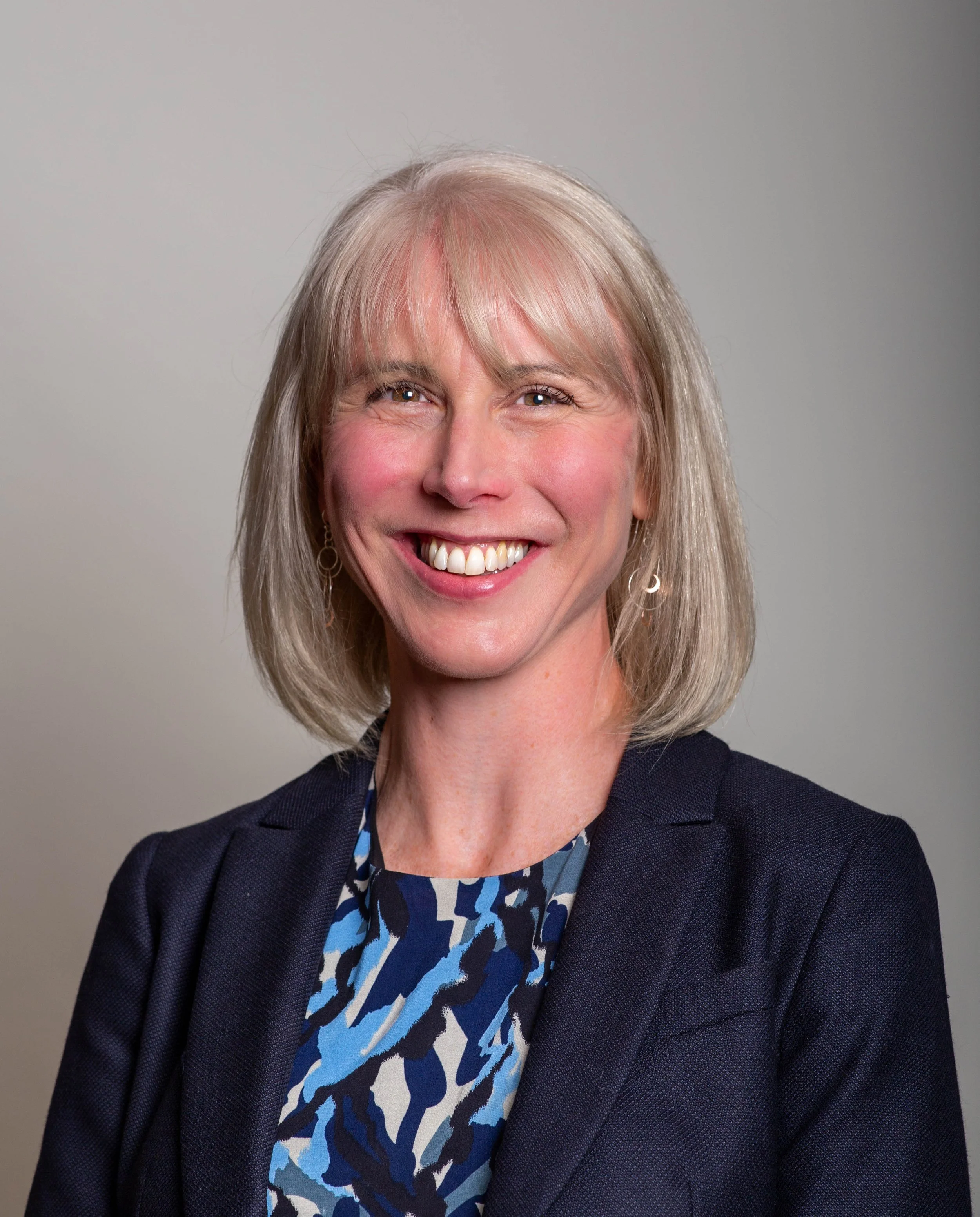 A woman with short silver hair smiling, wearing a patterned blue shirt and a dark blazer, against a plain background.
