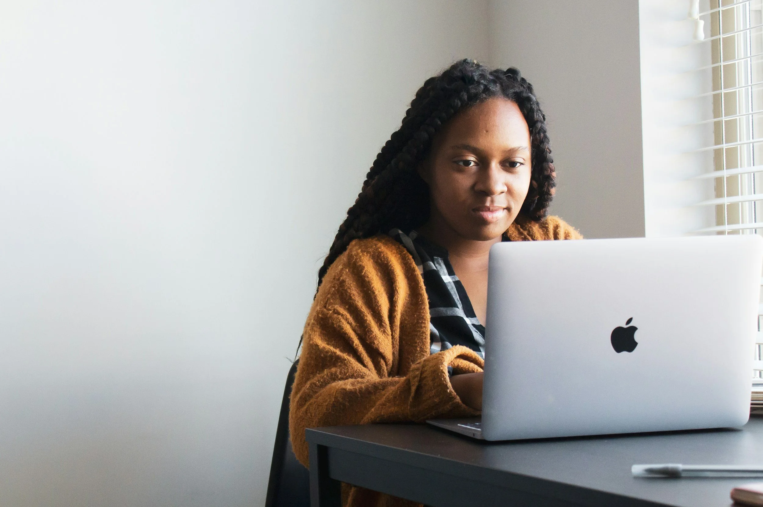 Young woman with braided hair working on a silver MacBook at a desk near a window with blinds.
