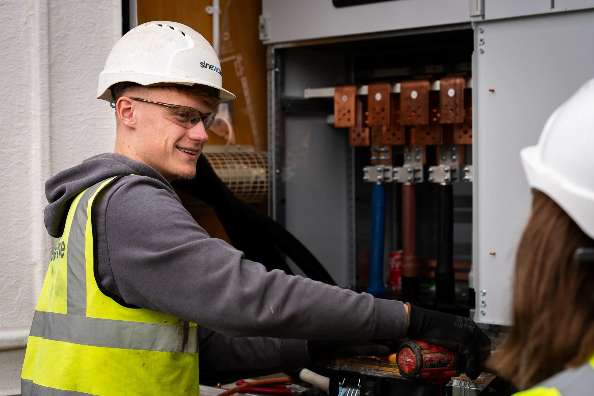 Electrician wearing a white hard hat and safety vest working inside an electrical panel.