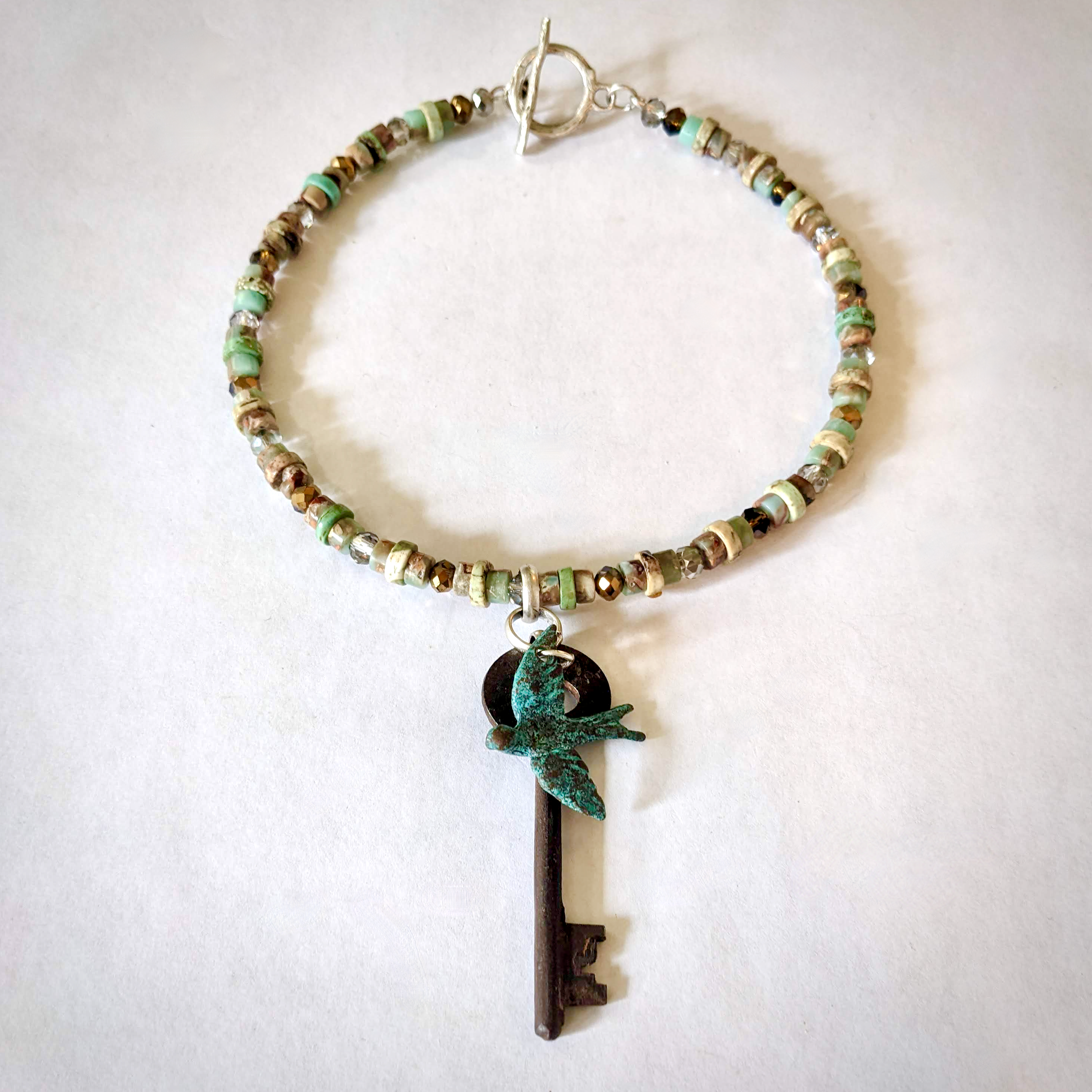 Antique iron key and verdigris swallow charm on choker-length silver jewelry wire strung with Amazonite and Imperial Jasper disc beads, and dove grey and amber faceted Czech glass beads.