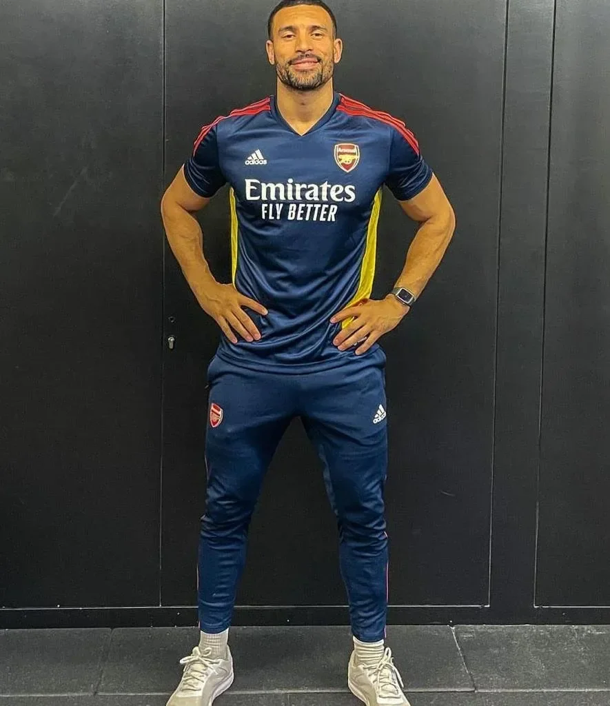 Man wearing a blue Arsenal football jersey with sponsor logos, standing with hands on hips against a black wall.