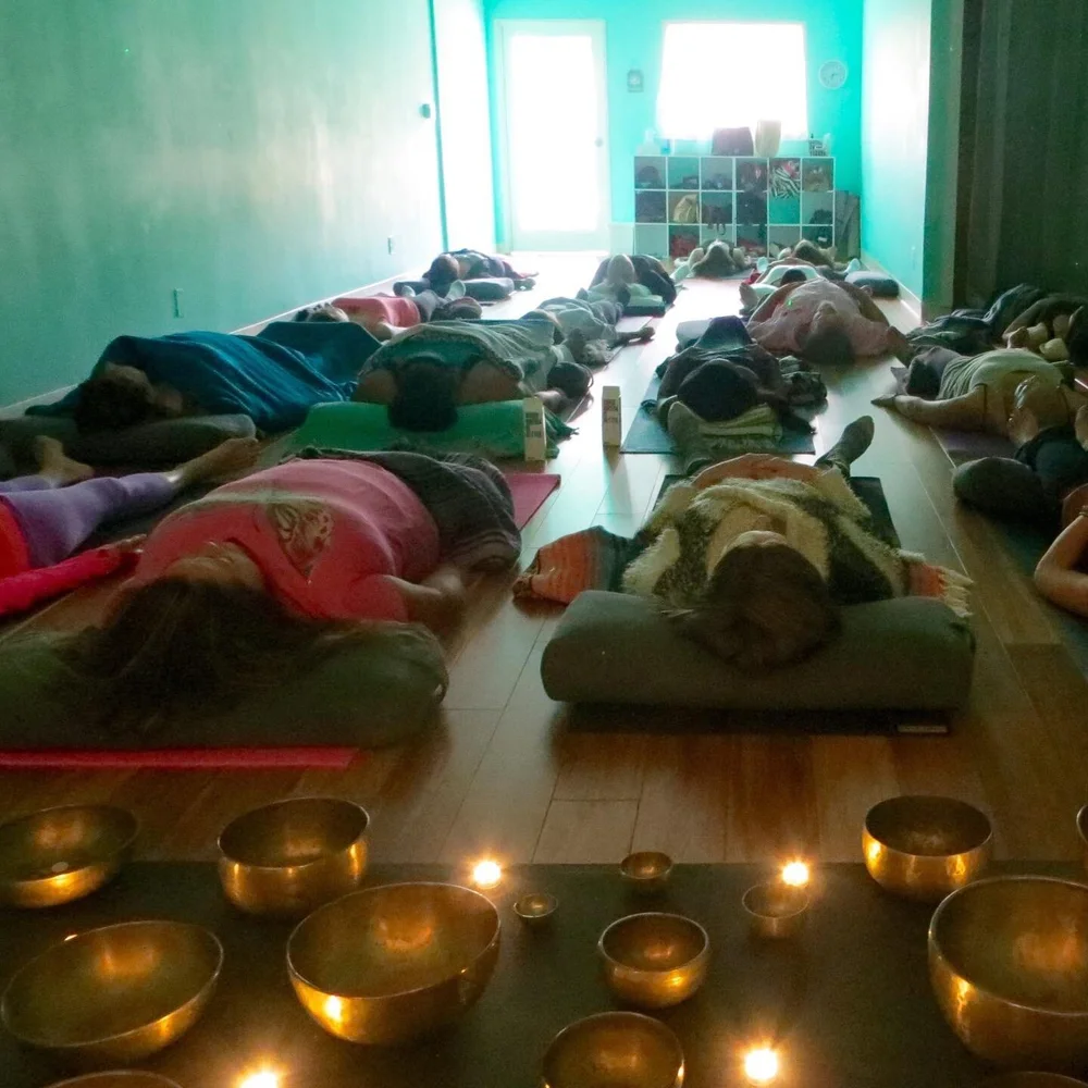 Students lying under blankets in rows during a candlelit sound bath, a line of brass singing bowls along the floor