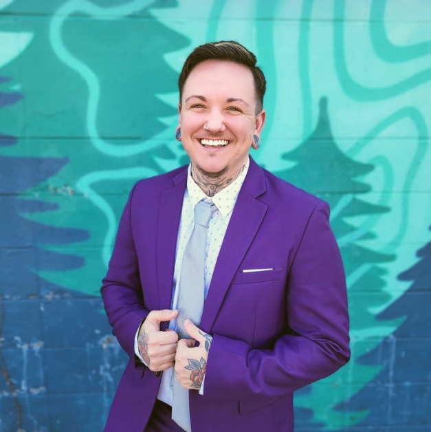 Person with short dark hair, tattoos on neck and hands, wearing a purple suit and light tie, smiling in front of teal and blue painted wall.