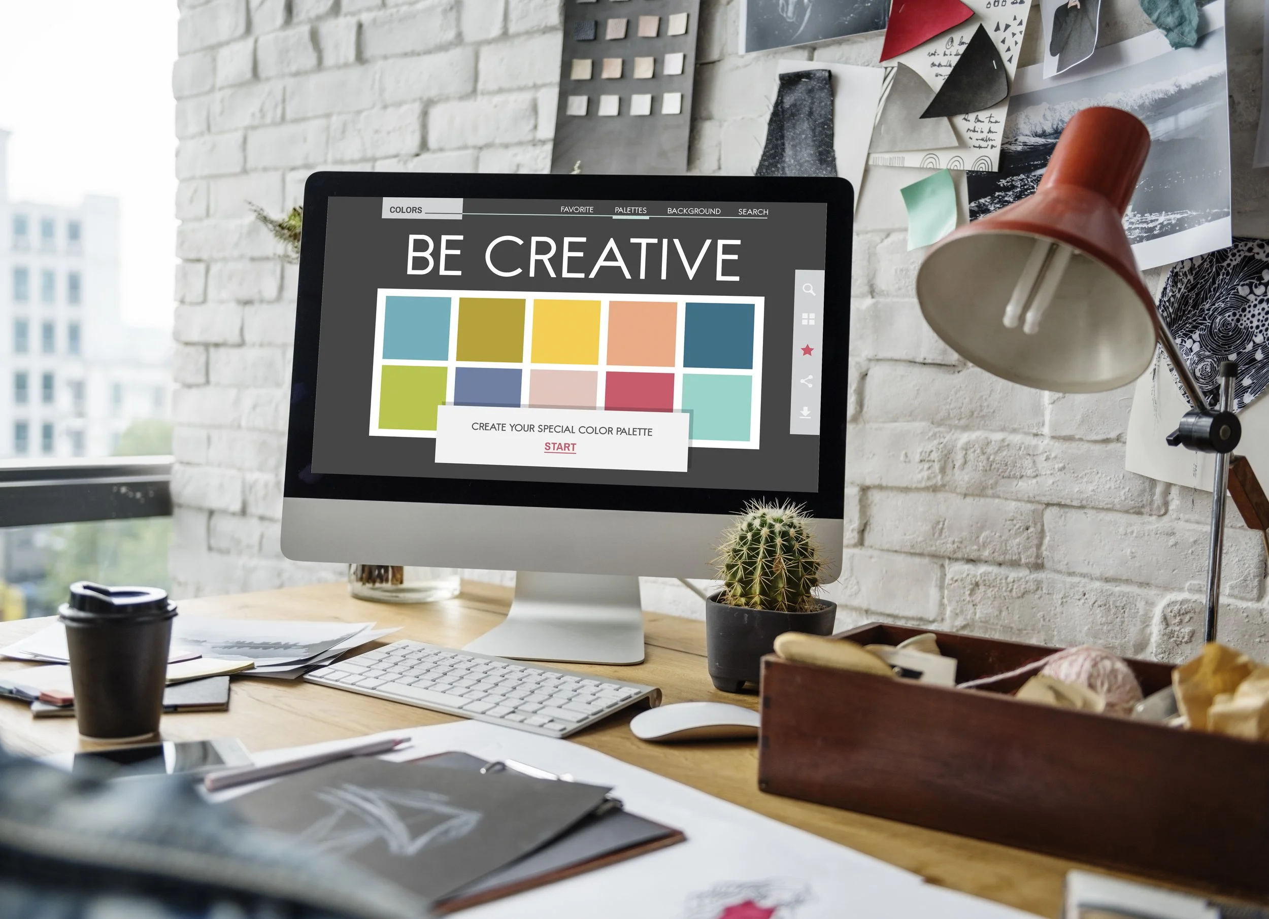 A creative workspace with a computer monitor displaying a colorful palette and the words "Be Creative." The desk has a cup of coffee, notebooks, and art supplies. The background features a white brick wall with pinned artwork and photos, a cactus, and a red desk lamp.